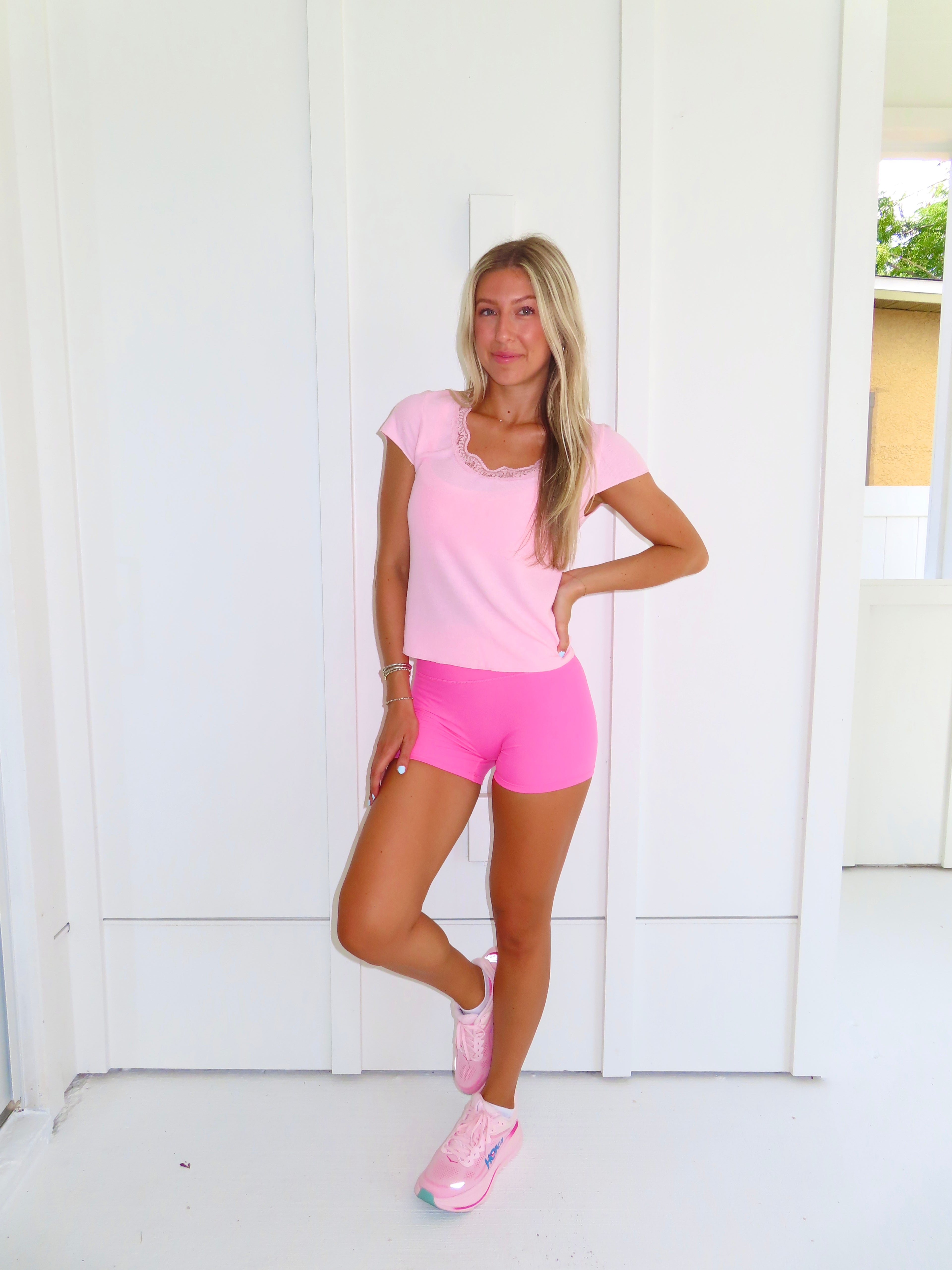 Woman wearing a pink tee and pink bikers shorts with pink sneakers standing against a white wall.
