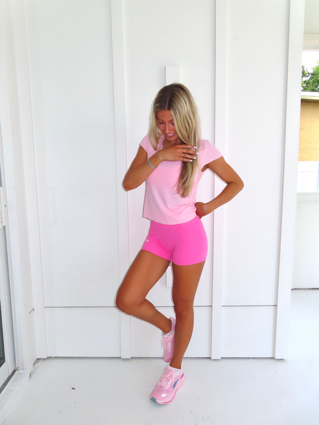 Woman wearing a pink tee and pink bikers shorts with pink sneakers standing against a white wall.