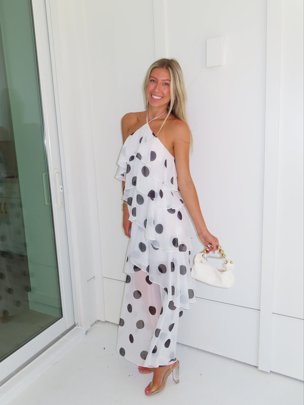 Woman wearing a white maxi dress with black polka dots holding a white purse and standing against a white wall.