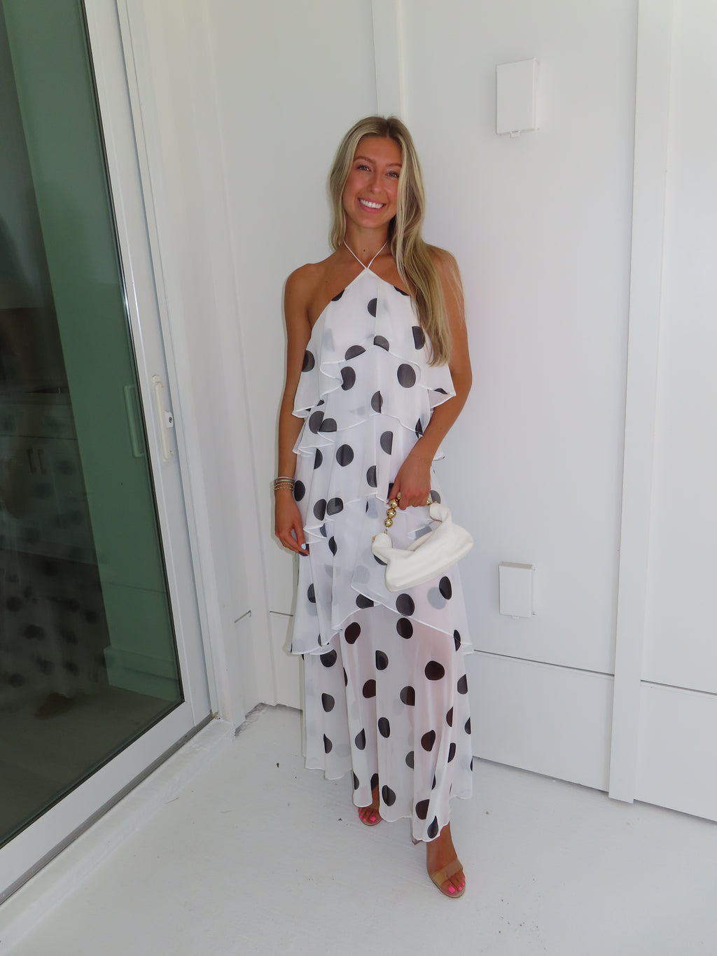 Woman wearing a white maxi dress with black polka dots holding a white purse and standing against a white wall.