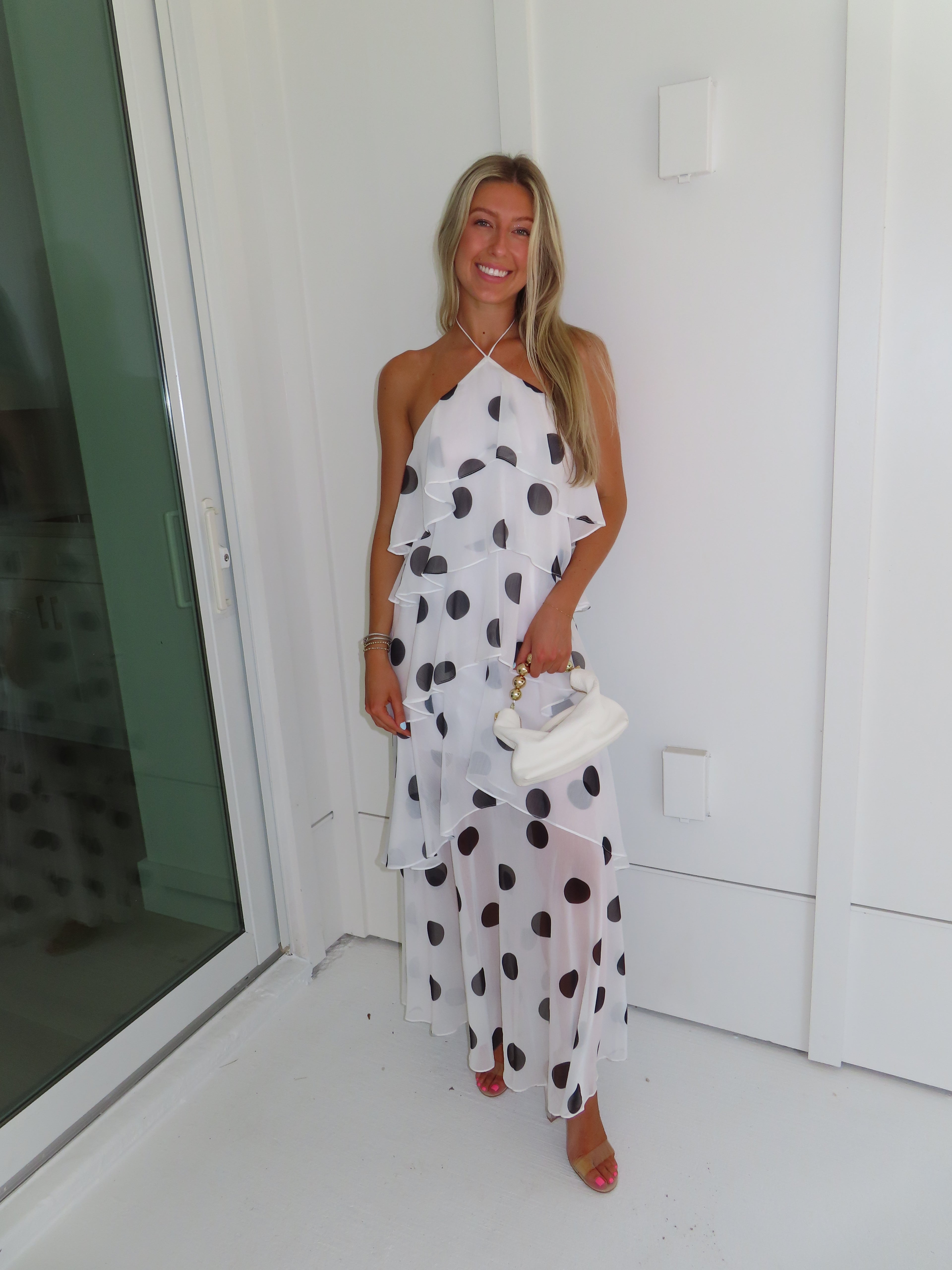 Woman wearing a white maxi dress with black polka dots holding a white purse and standing against a white wall.