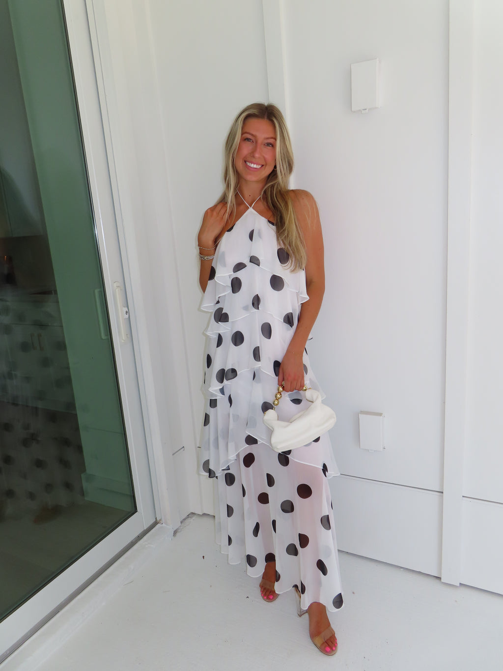 Woman wearing a white maxi dress with black polka dots holding a white purse and standing against a white wall.