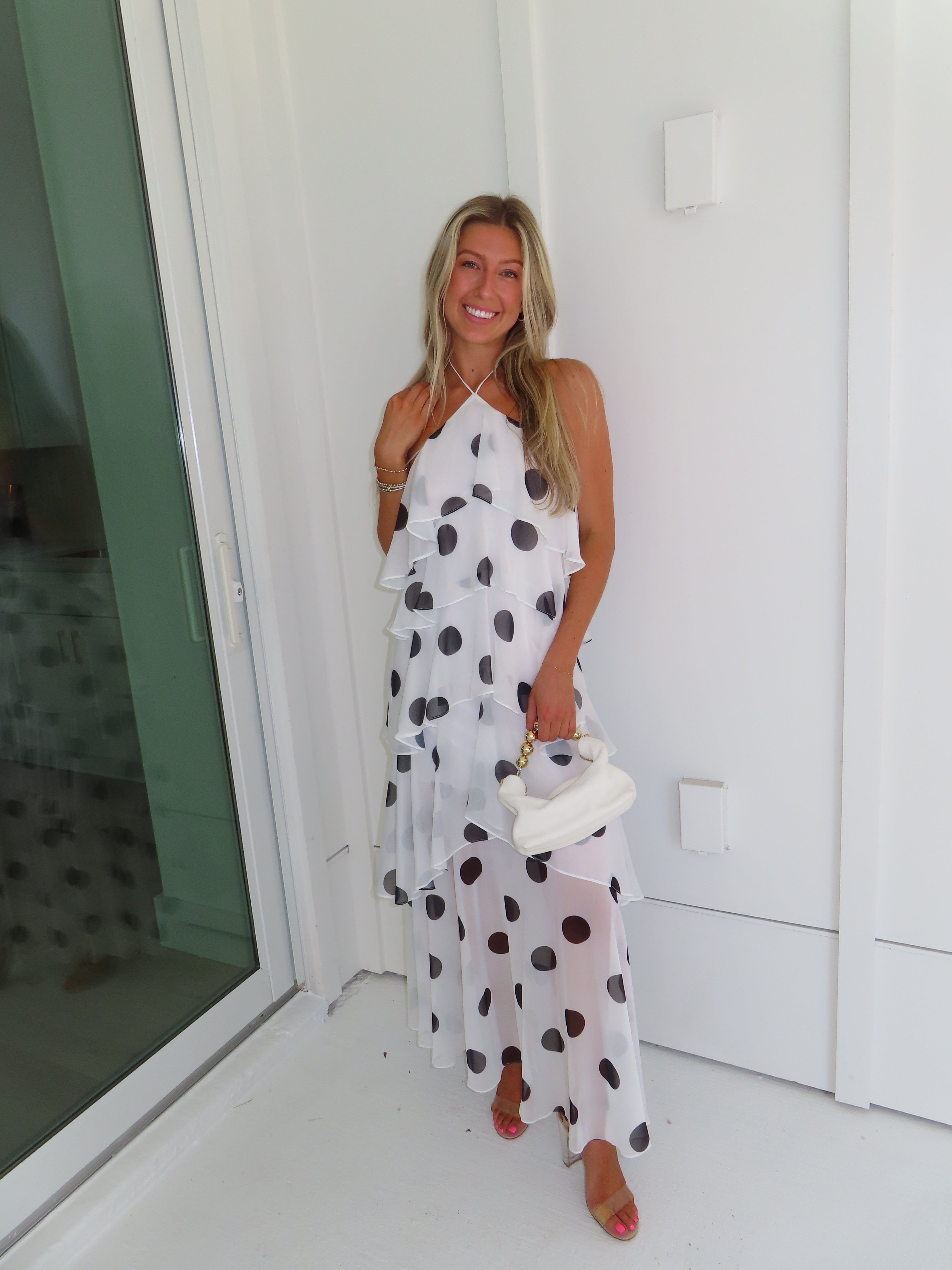 Woman wearing a white maxi dress with black polka dots holding a white purse and standing against a white wall.