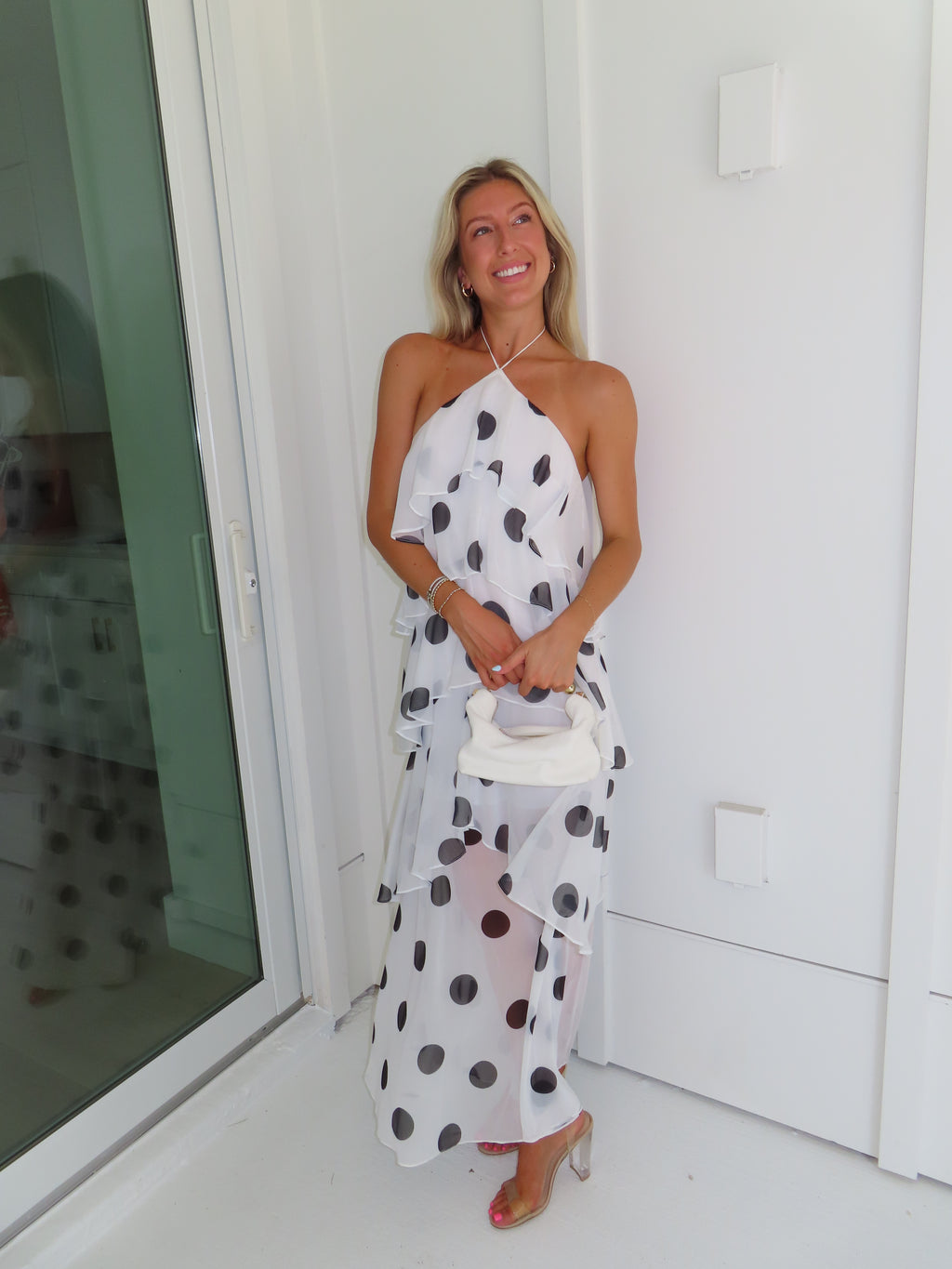 Woman wearing a white maxi dress with black polka dots holding a white purse and standing against a white wall.