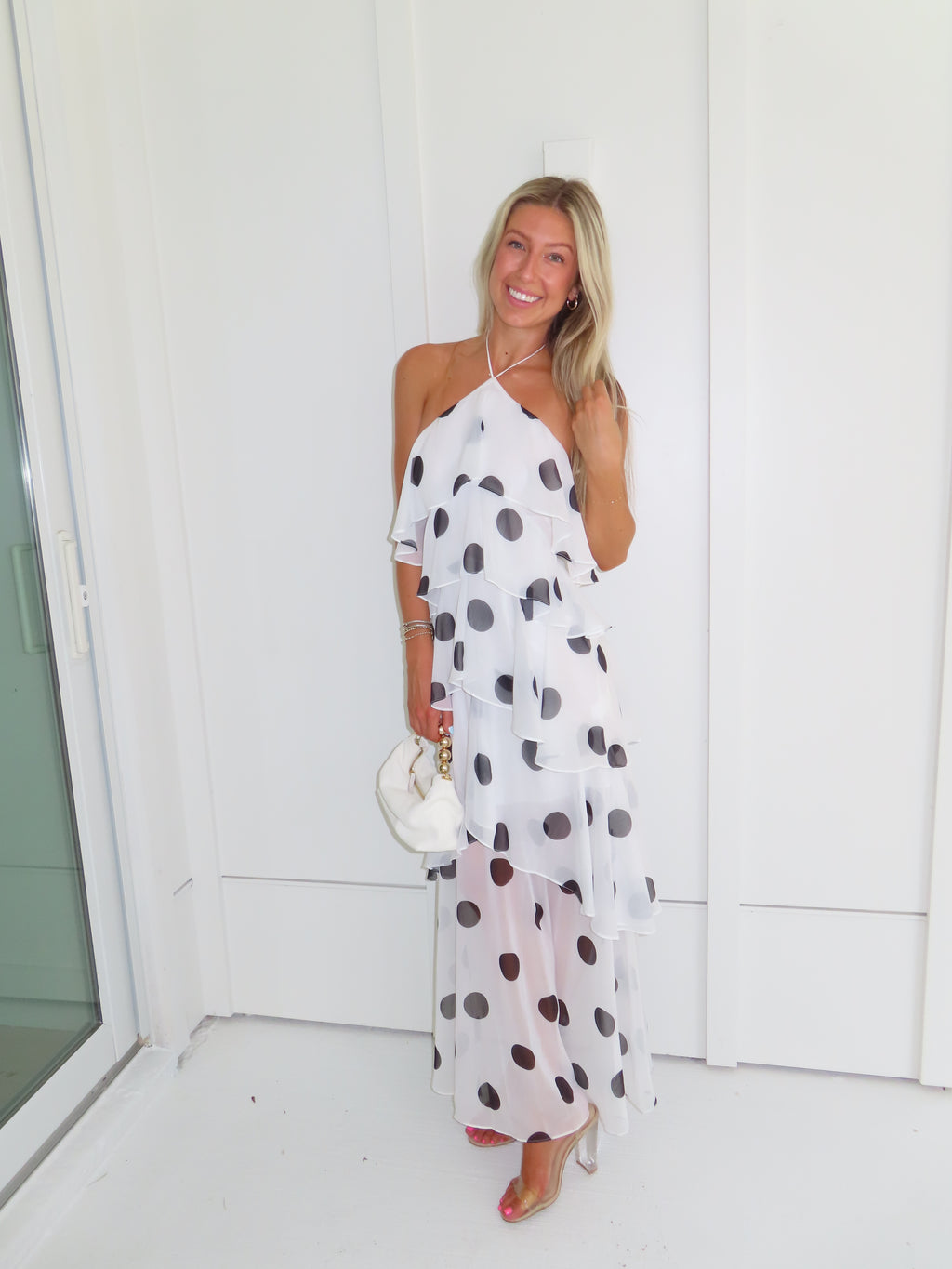 Woman wearing a white maxi dress with black polka dots holding a white purse and standing against a white wall.