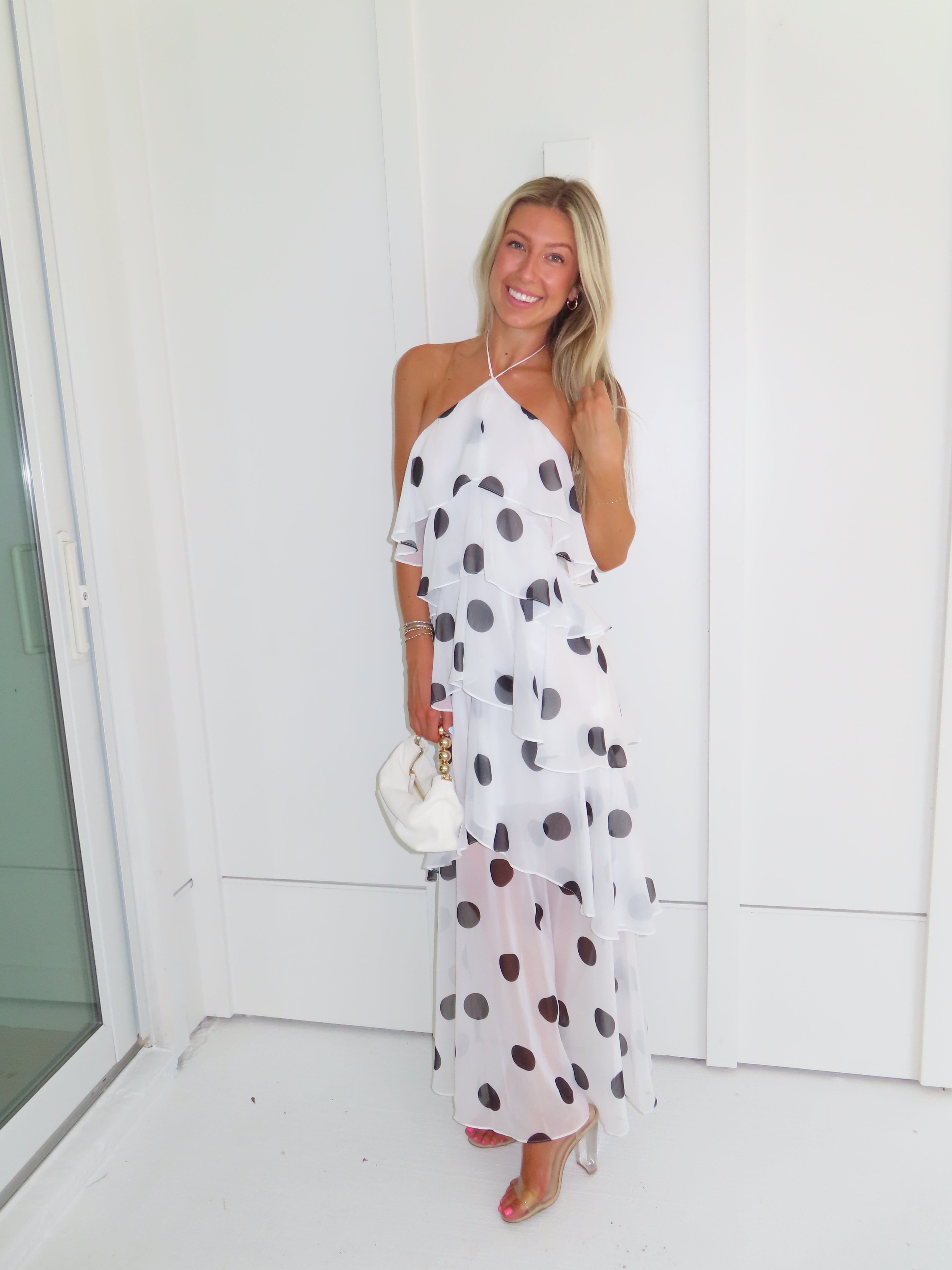 Woman wearing a white maxi dress with black polka dots holding a white purse and standing against a white wall.