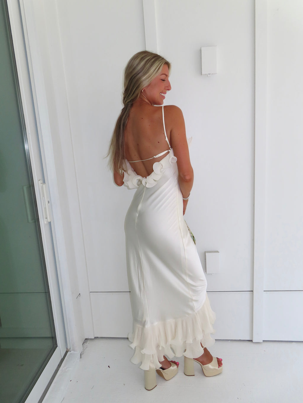 Woman wearing an ivory maxi dress with gold heels holding a green clutch against a white background.