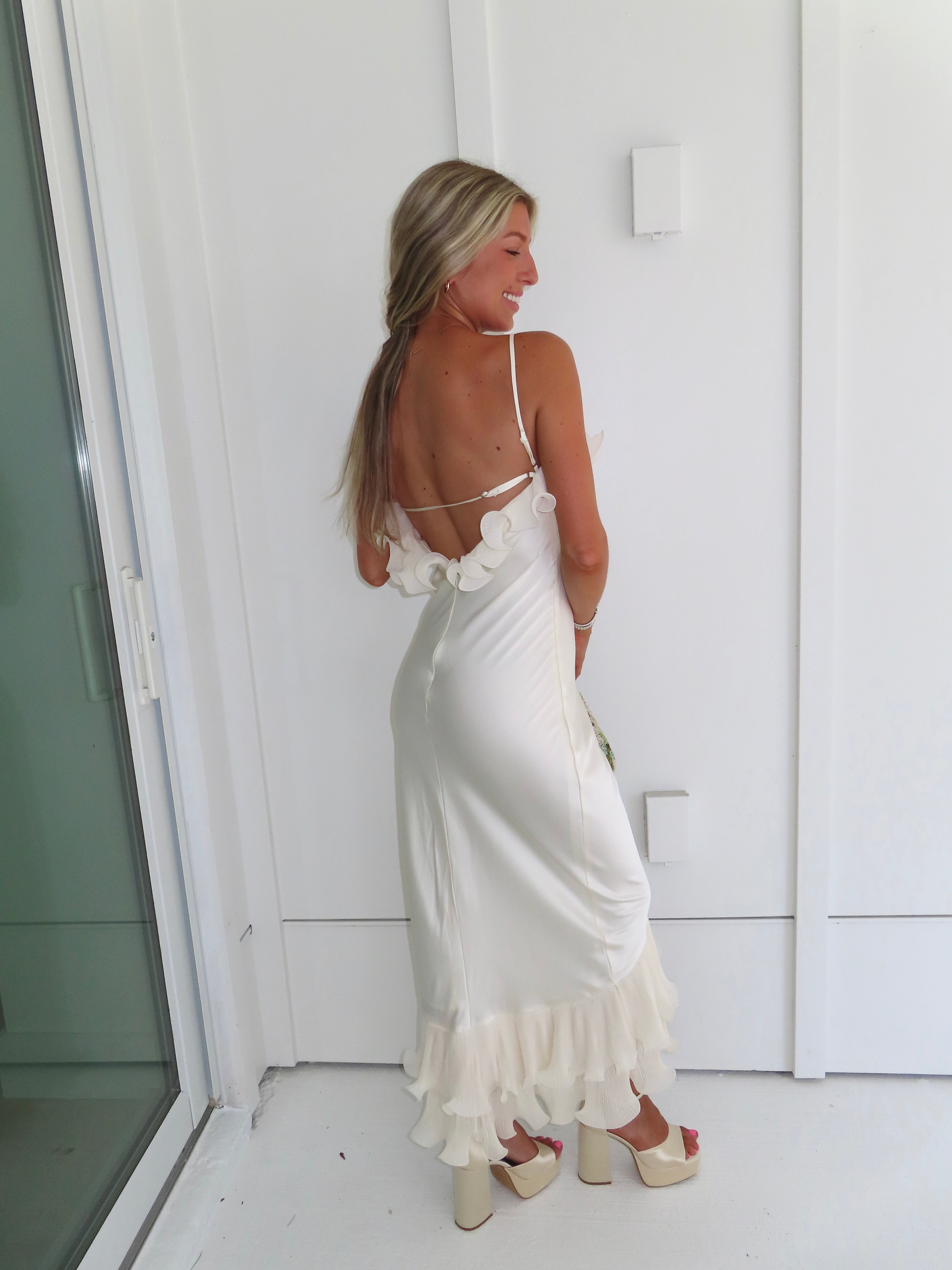 Woman wearing an ivory maxi dress with gold heels holding a green clutch against a white background.