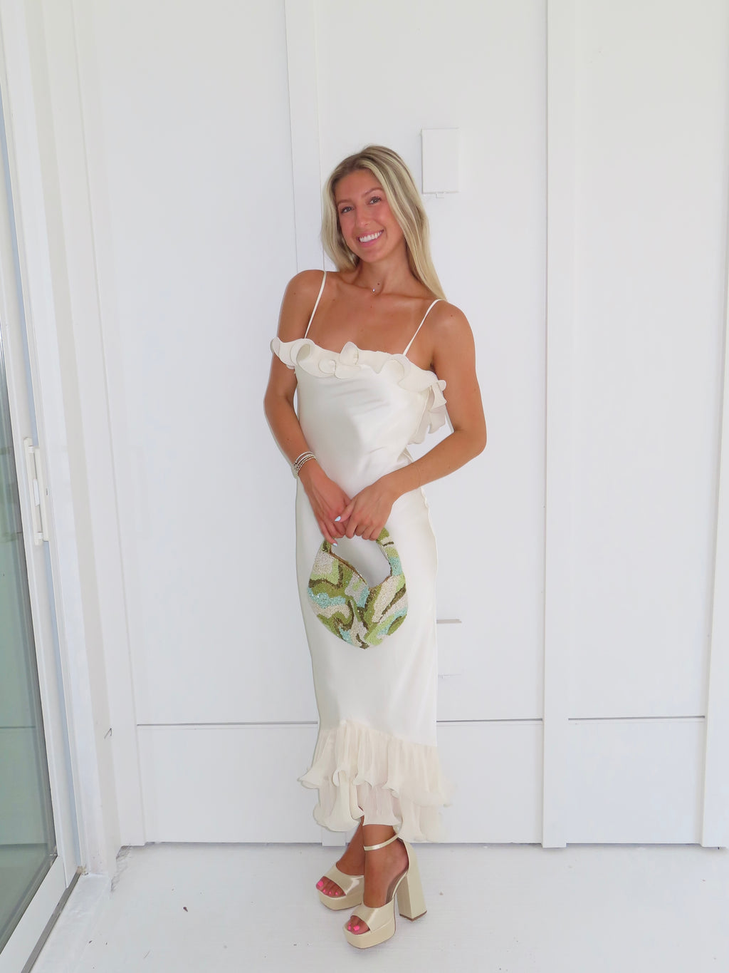 Woman wearing an ivory maxi dress with gold heels holding a green clutch against a white background.