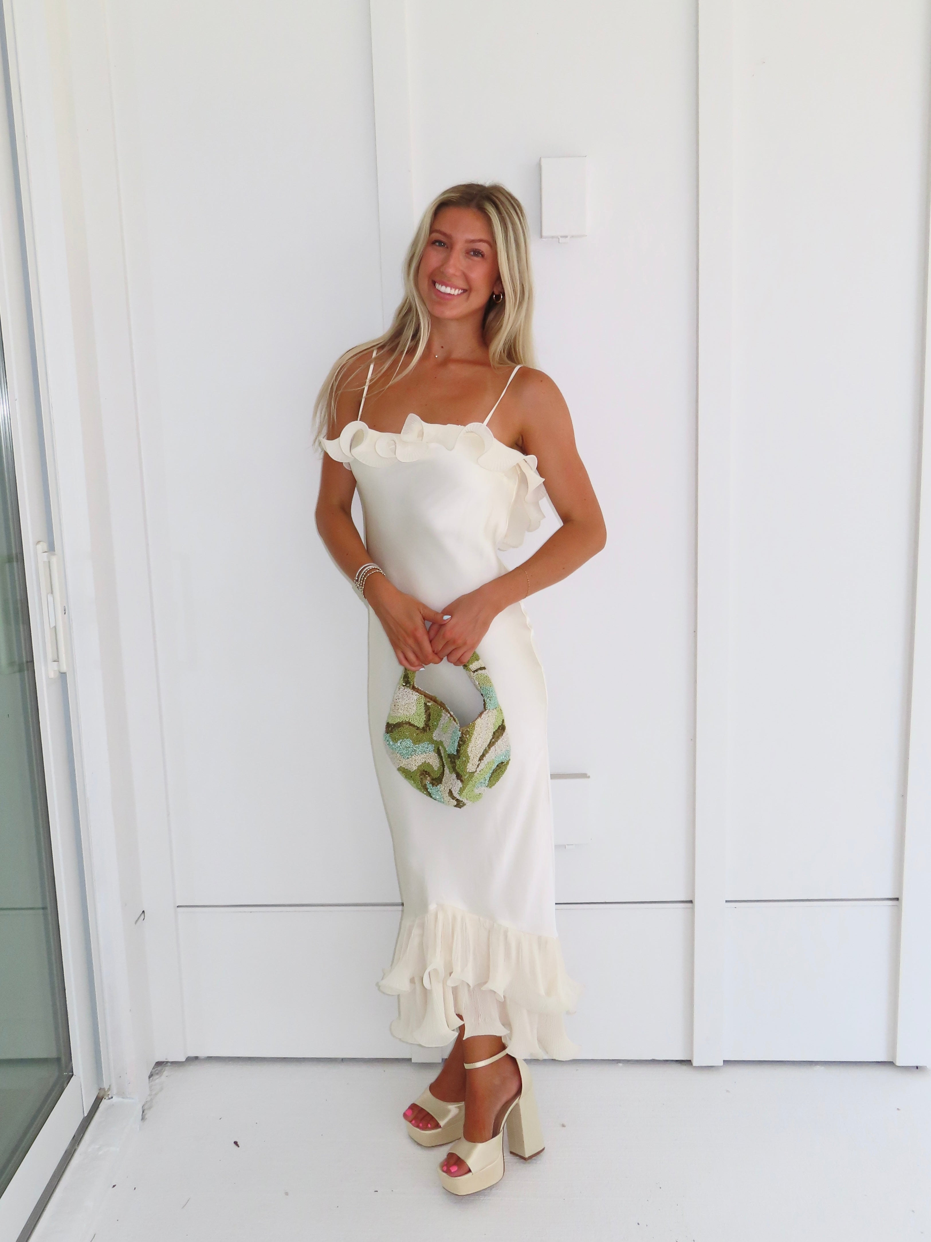 Woman wearing an ivory maxi dress with gold heels holding a green clutch against a white background.