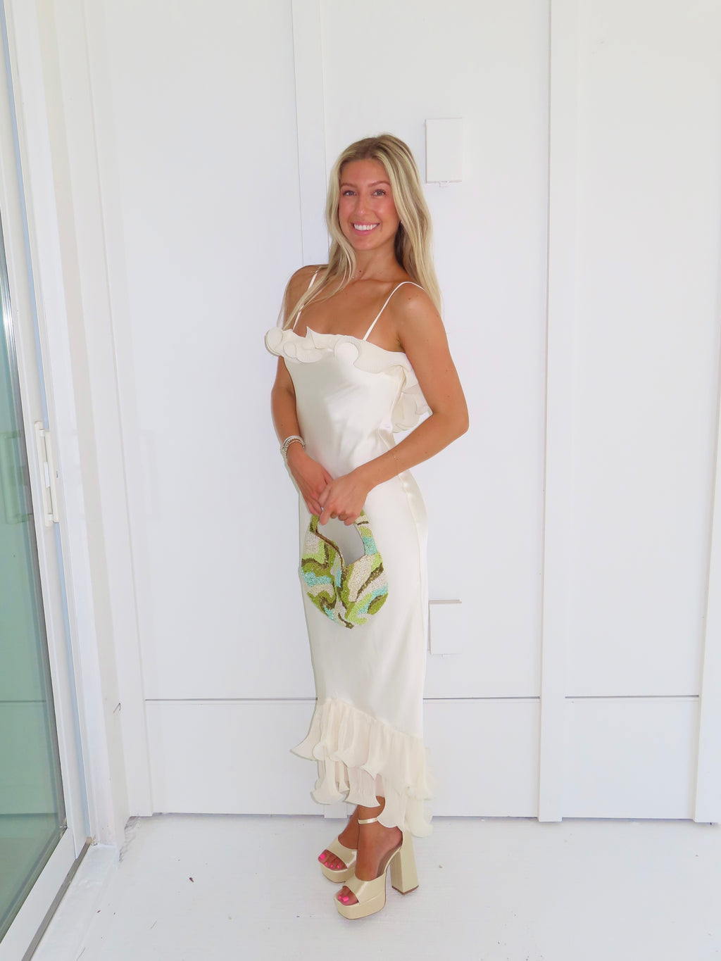 Woman wearing an ivory maxi dress with gold heels holding a green clutch against a white background.