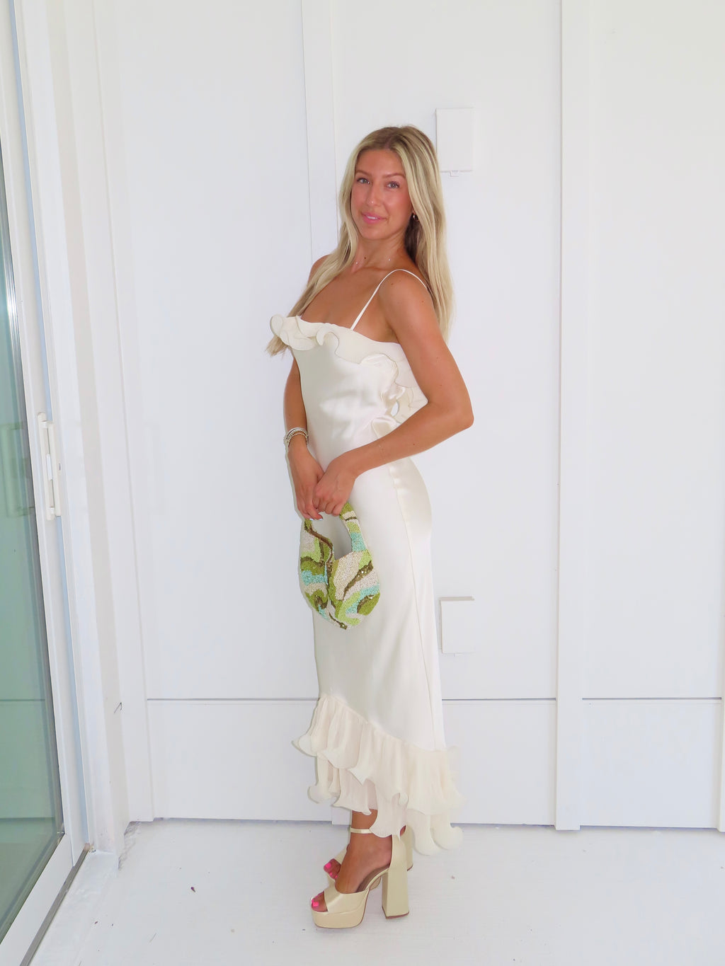 Woman wearing an ivory maxi dress with gold heels holding a green clutch against a white background.