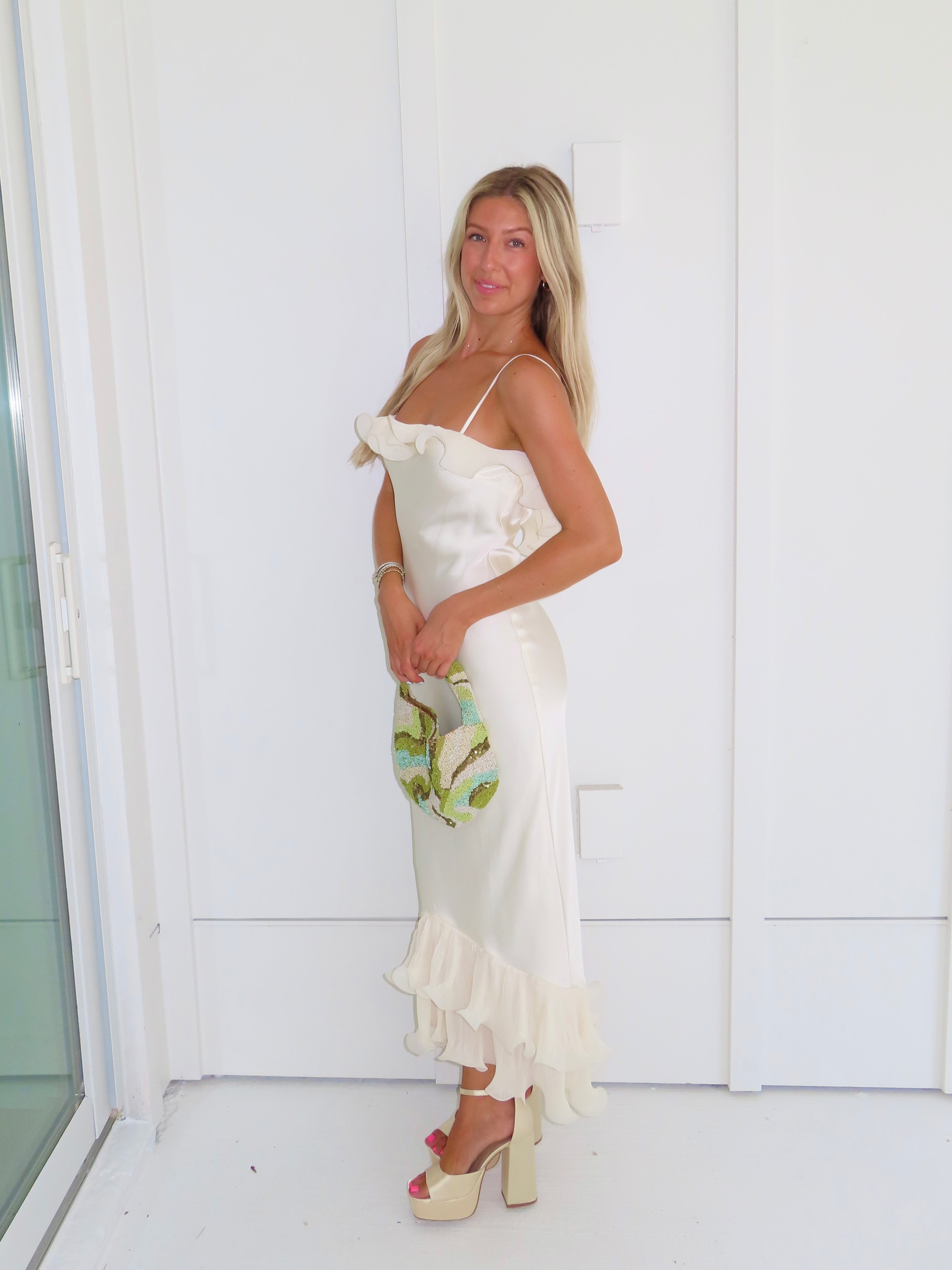 Woman wearing an ivory maxi dress with gold heels holding a green clutch against a white background.