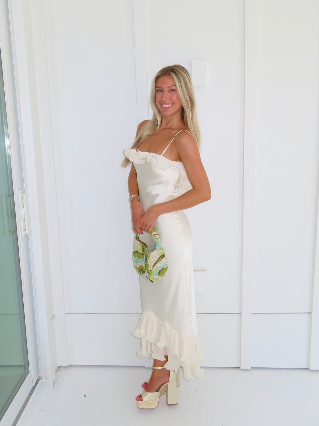 Woman wearing an ivory maxi dress with gold heels holding a green clutch against a white background.