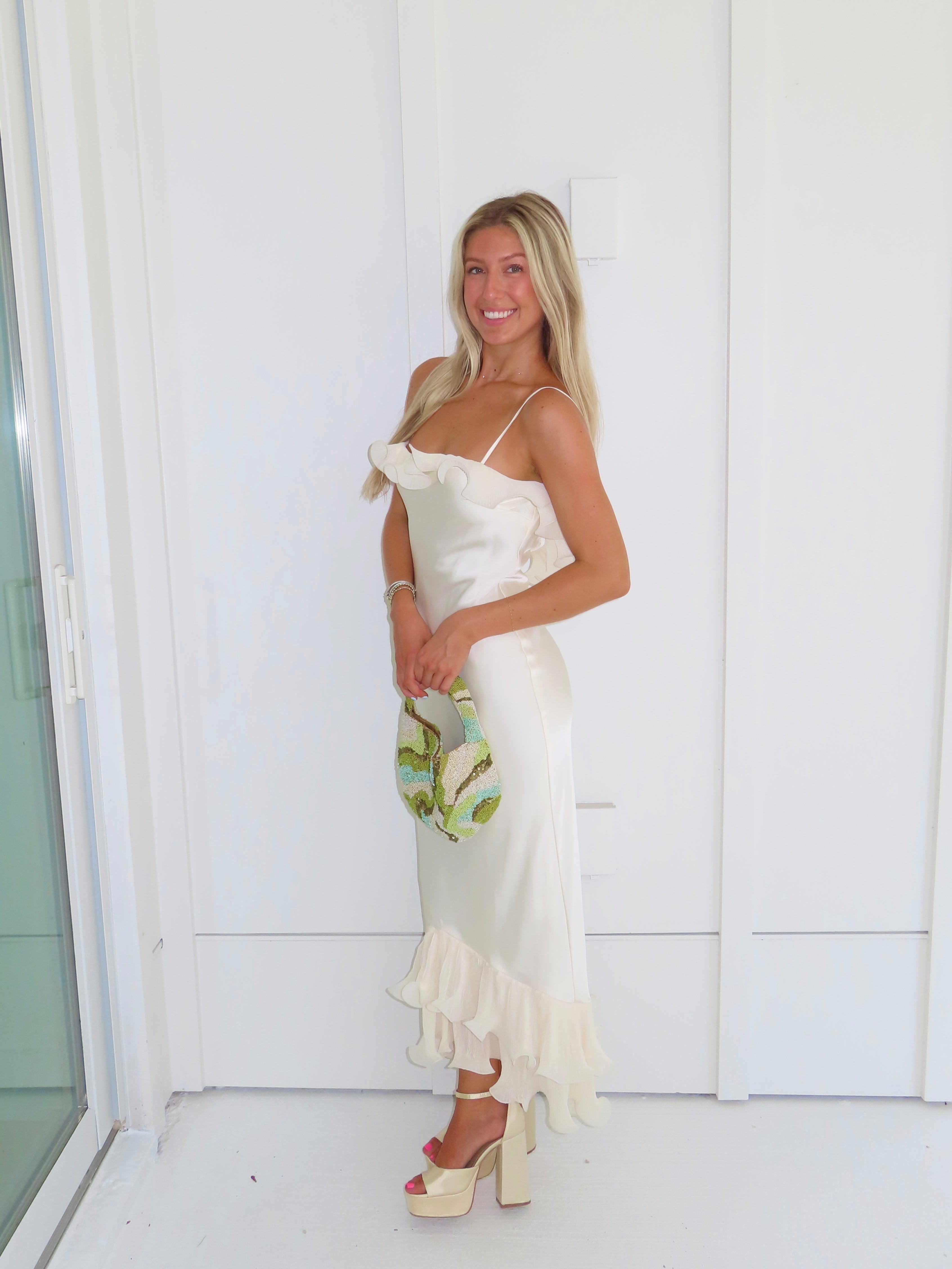 Woman wearing an ivory maxi dress with gold heels holding a green clutch against a white background.
