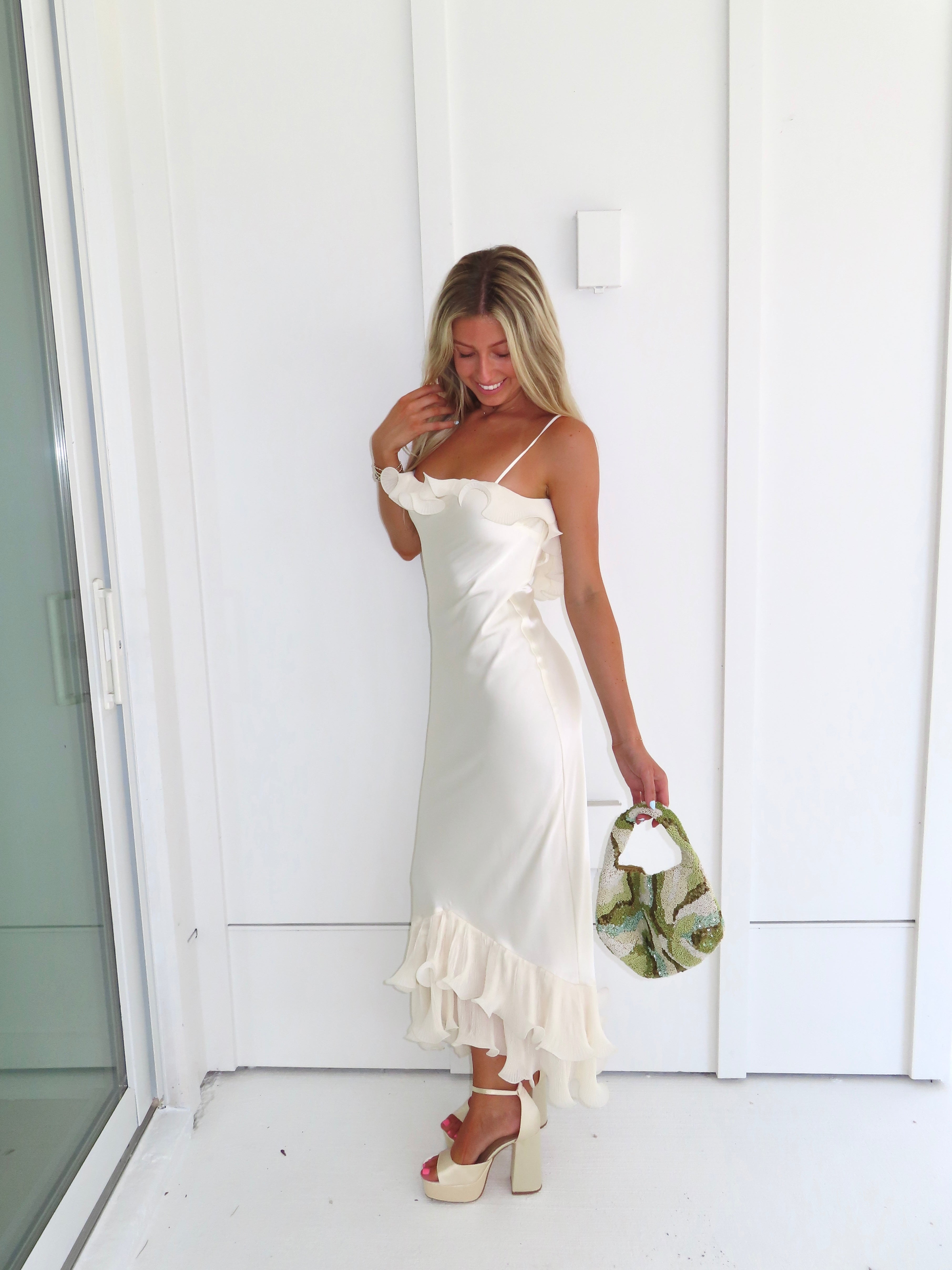Woman wearing an ivory maxi dress with gold heels holding a green clutch against a white background.