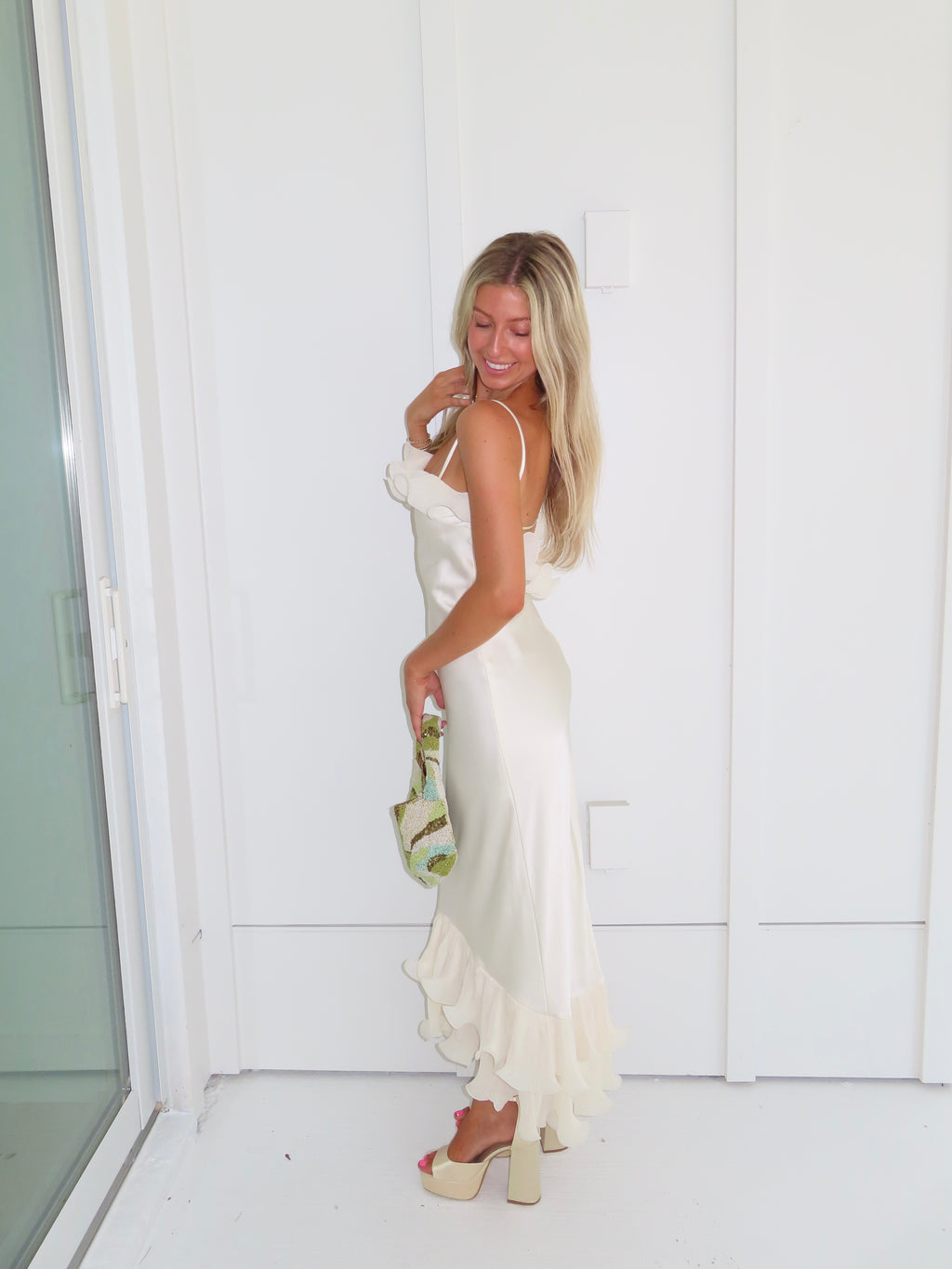 Woman wearing an ivory maxi dress with gold heels holding a green clutch against a white background.