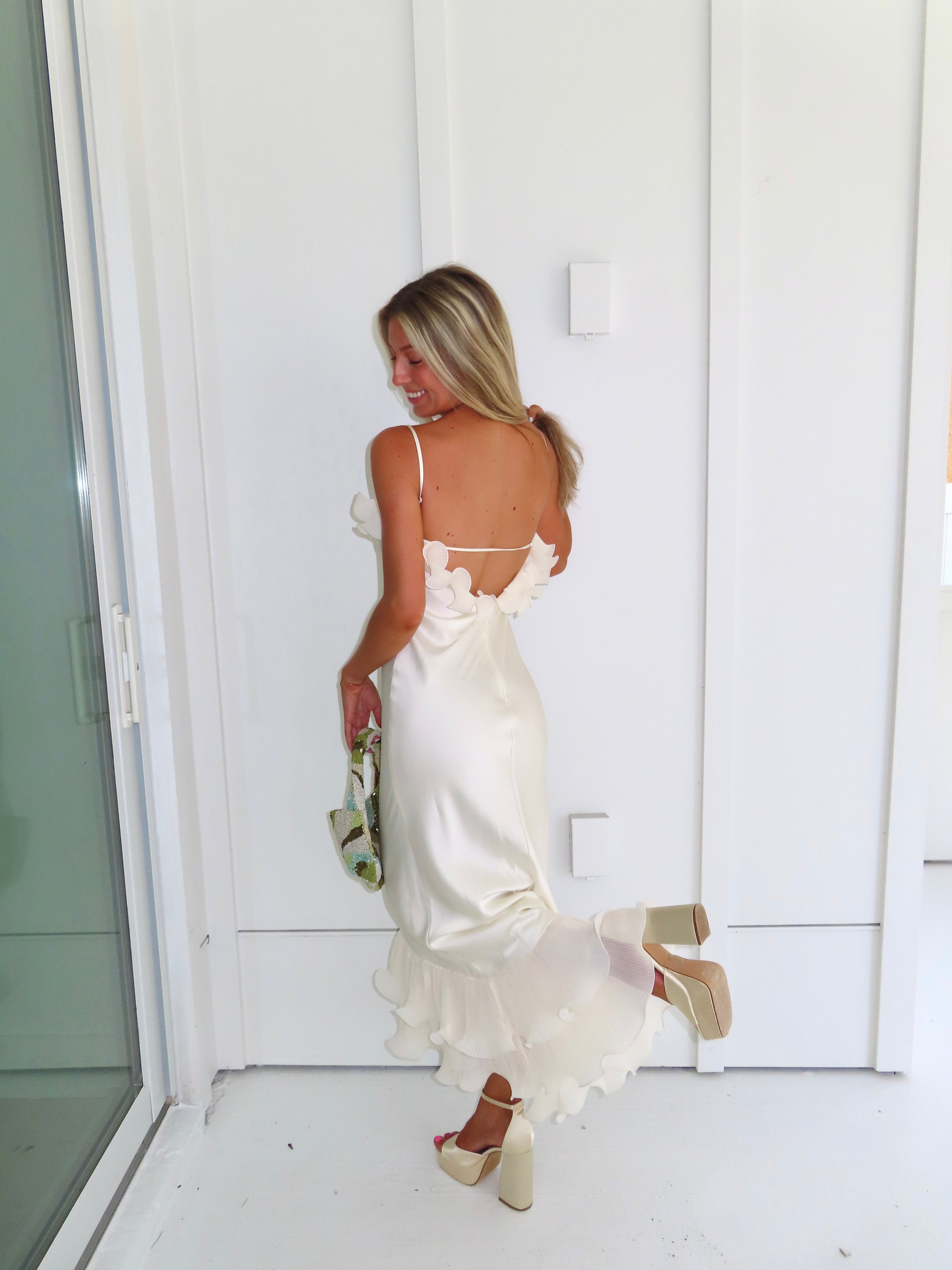 Woman wearing an ivory maxi dress with gold heels holding a green clutch against a white background.