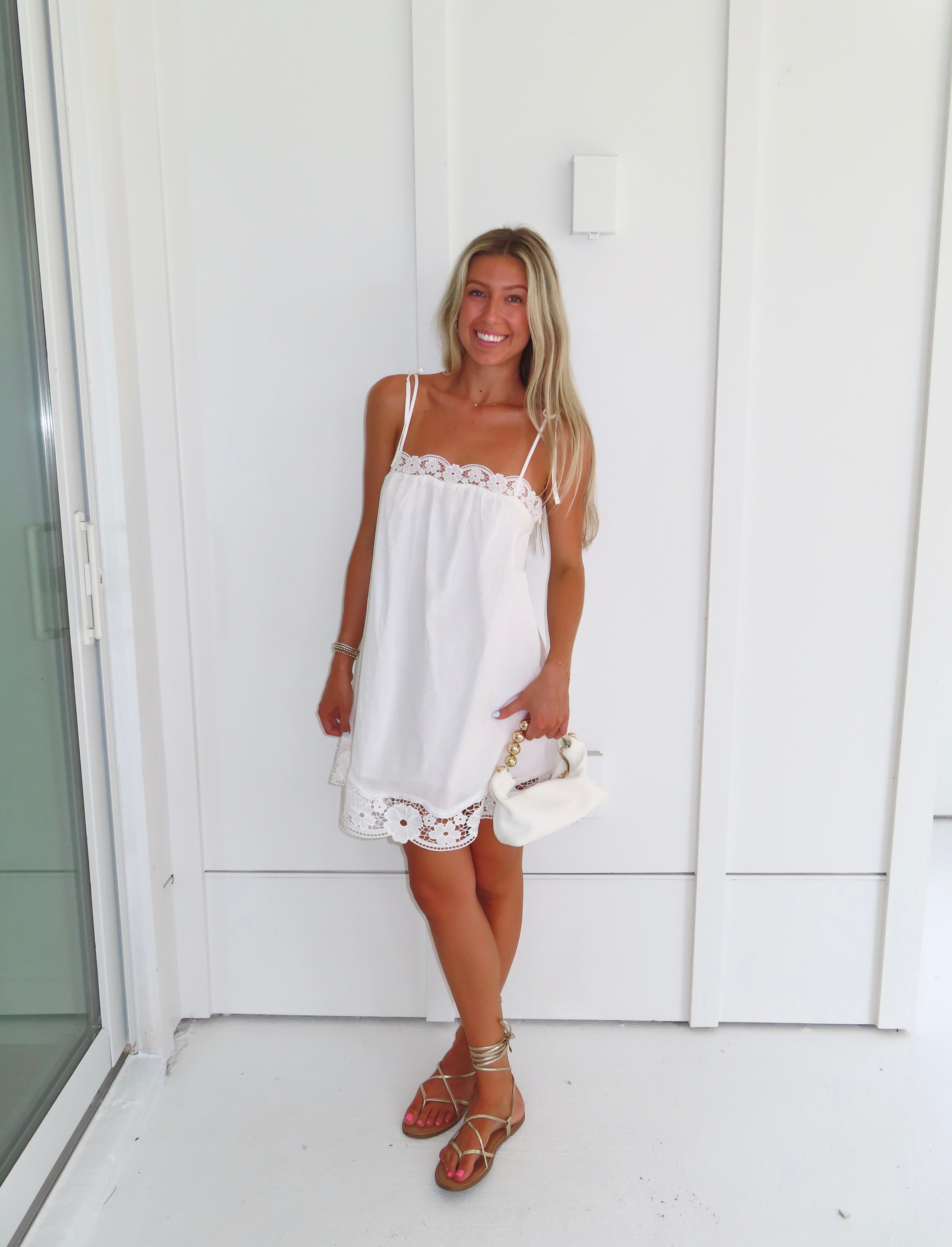 Woman wearing a white dress with lace details and gold sandals holding a white purse against a white wall.