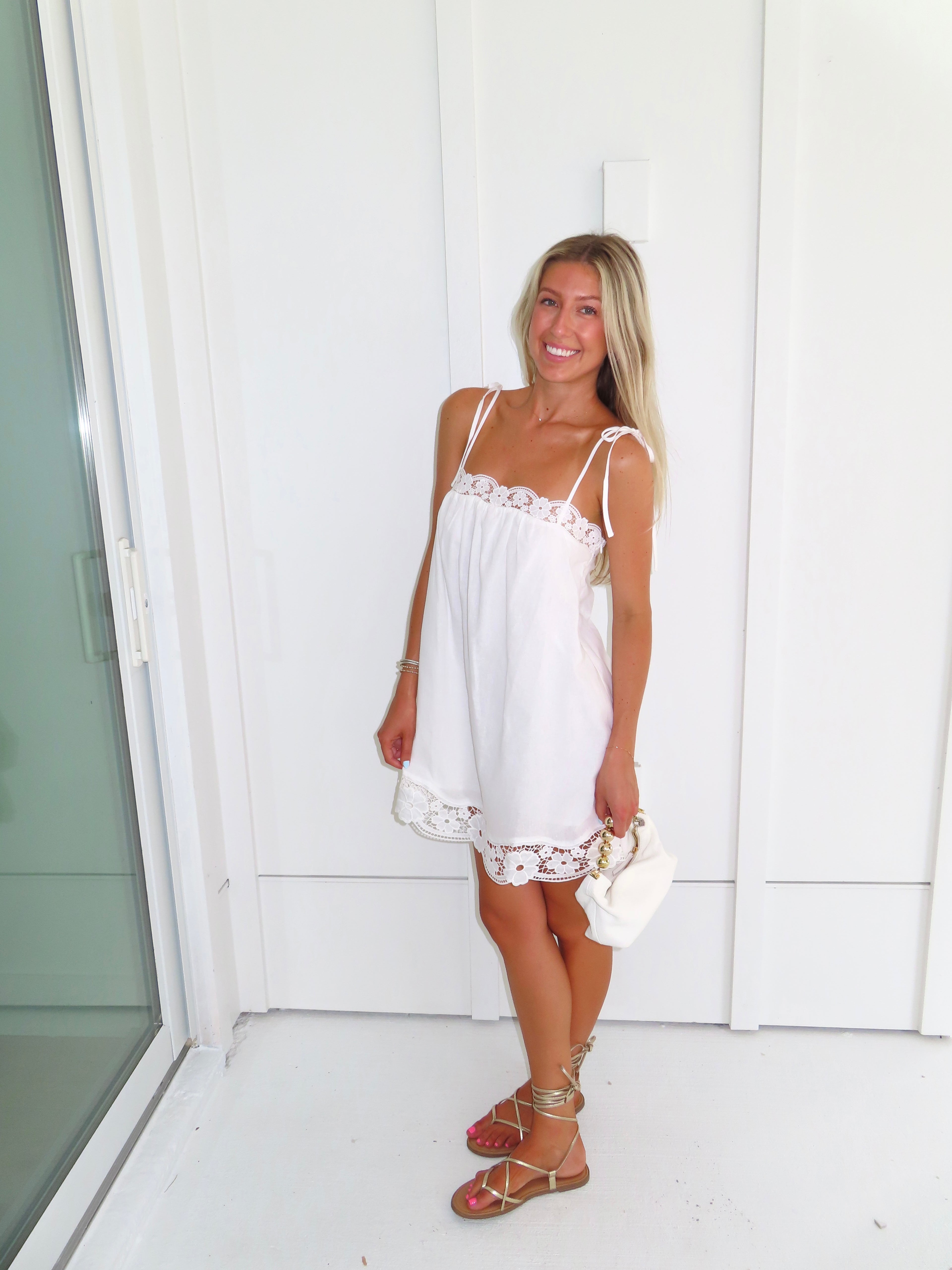 Woman wearing a white dress with lace details and gold sandals holding a white purse against a white wall.