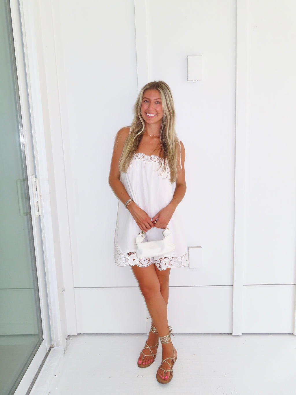 Woman wearing a white dress with lace details and gold sandals holding a white purse against a white wall.