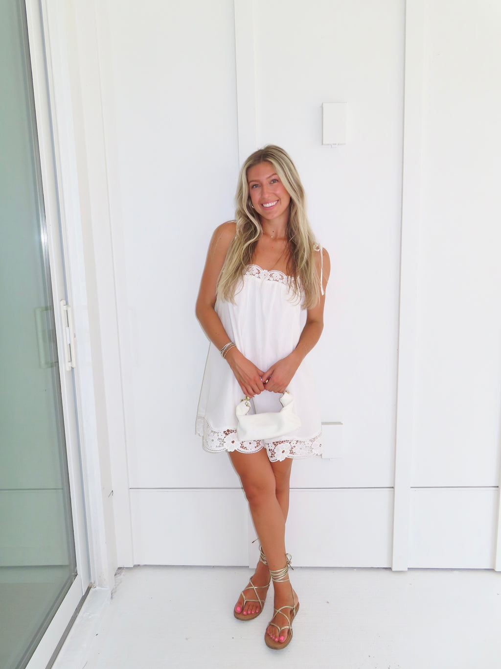 Woman wearing a white dress with lace details and gold sandals holding a white purse against a white wall.
