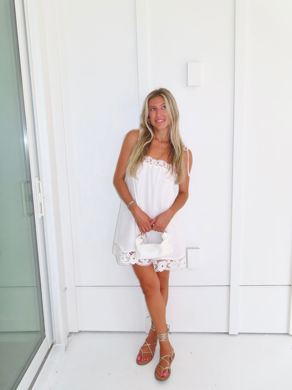Woman wearing a white dress with lace details and gold sandals holding a white purse against a white wall.