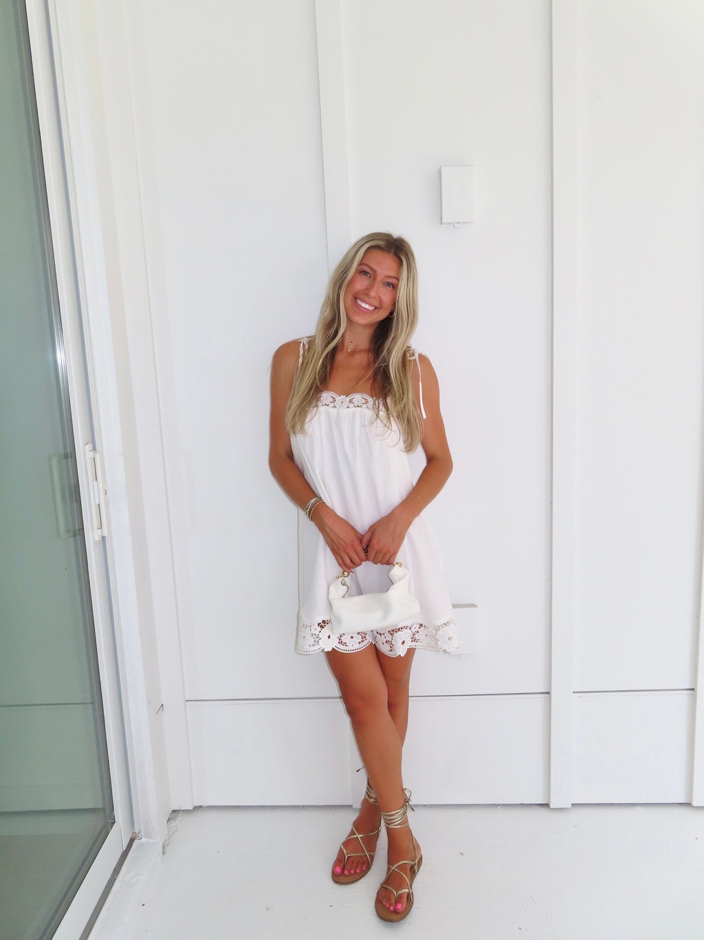 Woman wearing a white dress with lace details and gold sandals holding a white purse against a white wall.