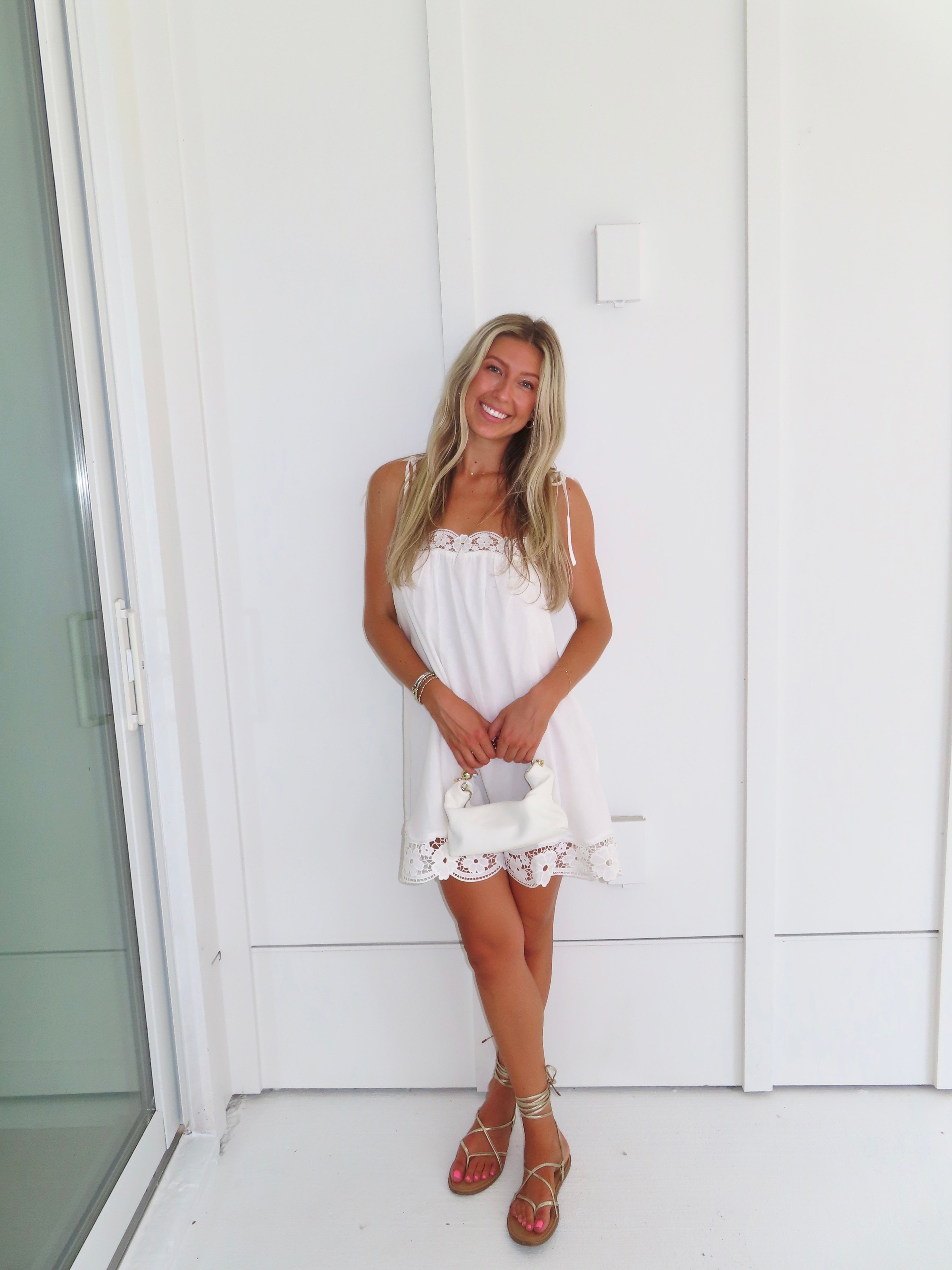 Woman wearing a white dress with lace details and gold sandals holding a white purse against a white wall.