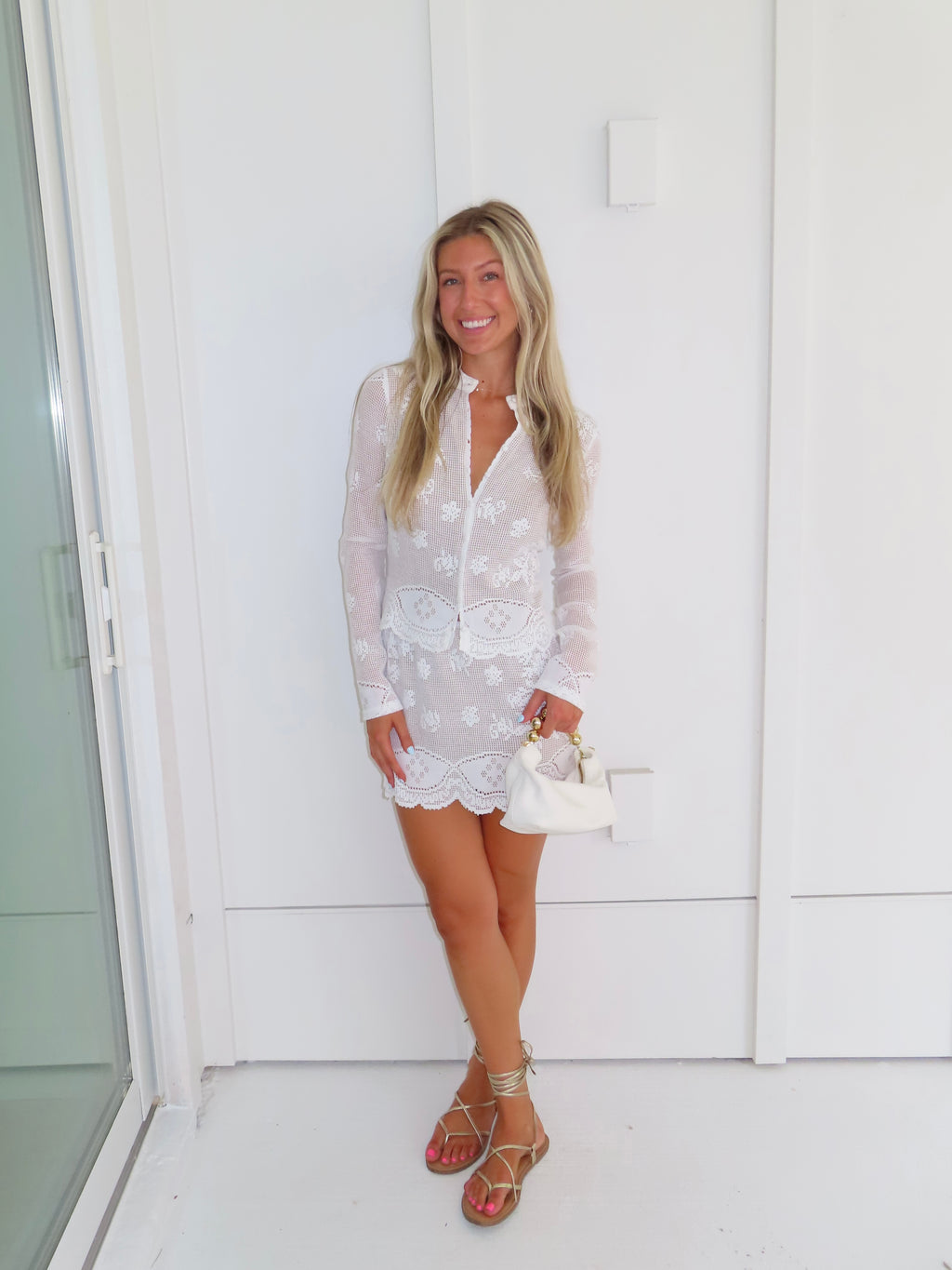 Woman wearing a white lace long sleeve and skirt set holding a white purse against a white wall.