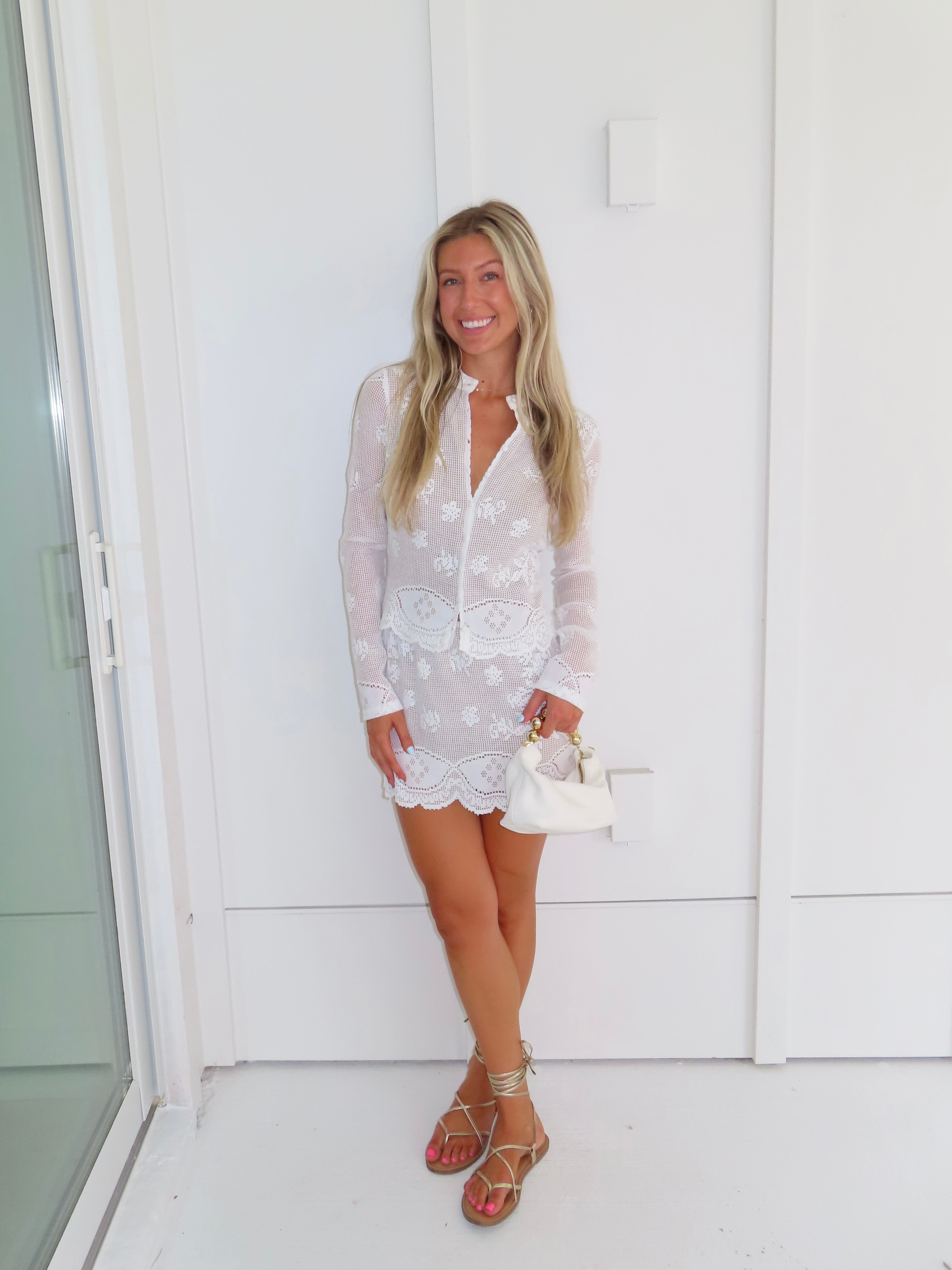 Woman wearing a white lace long sleeve and skirt set holding a white purse against a white wall.