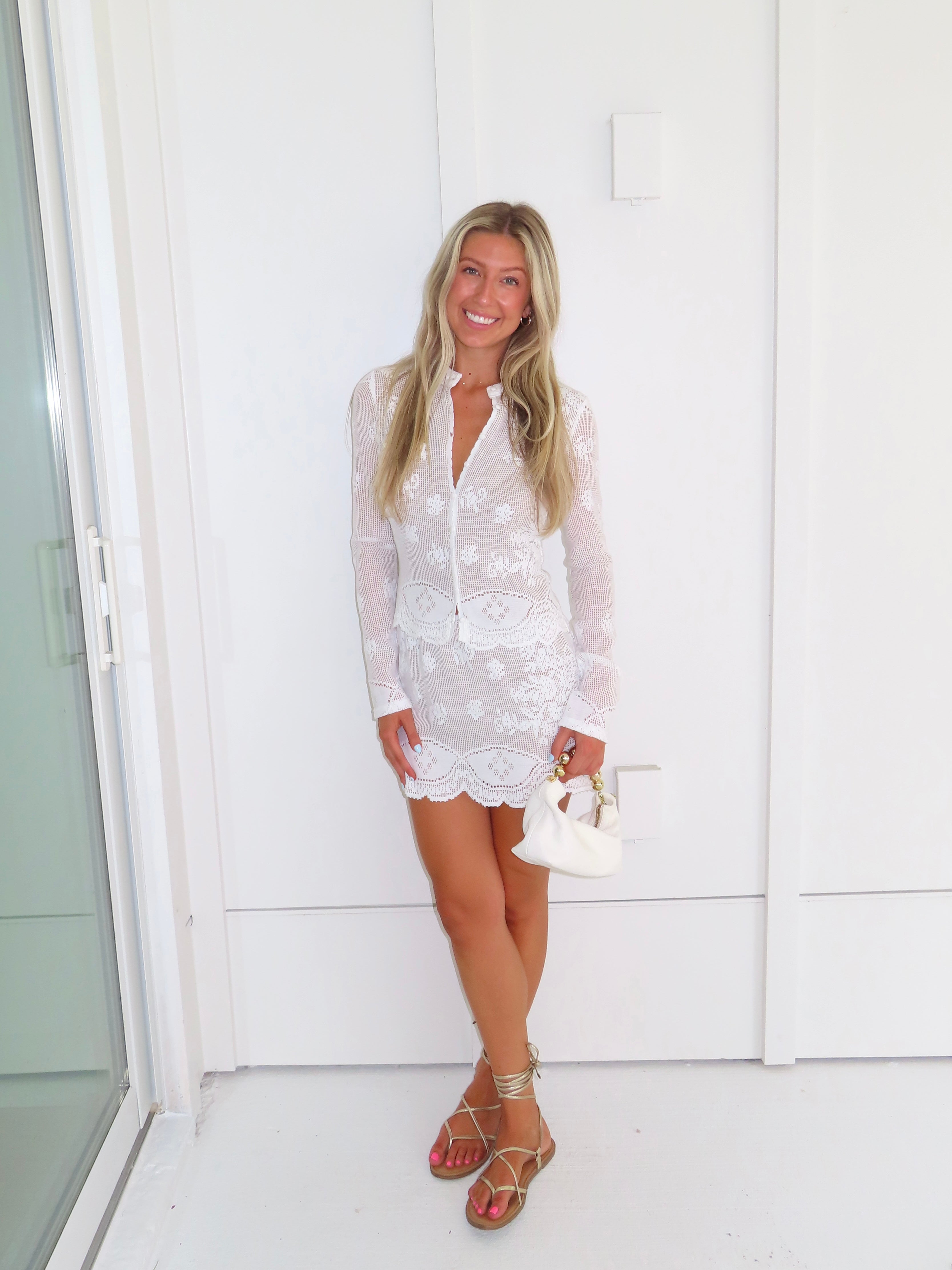 Woman wearing a white lace long sleeve and skirt set holding a white purse against a white wall.