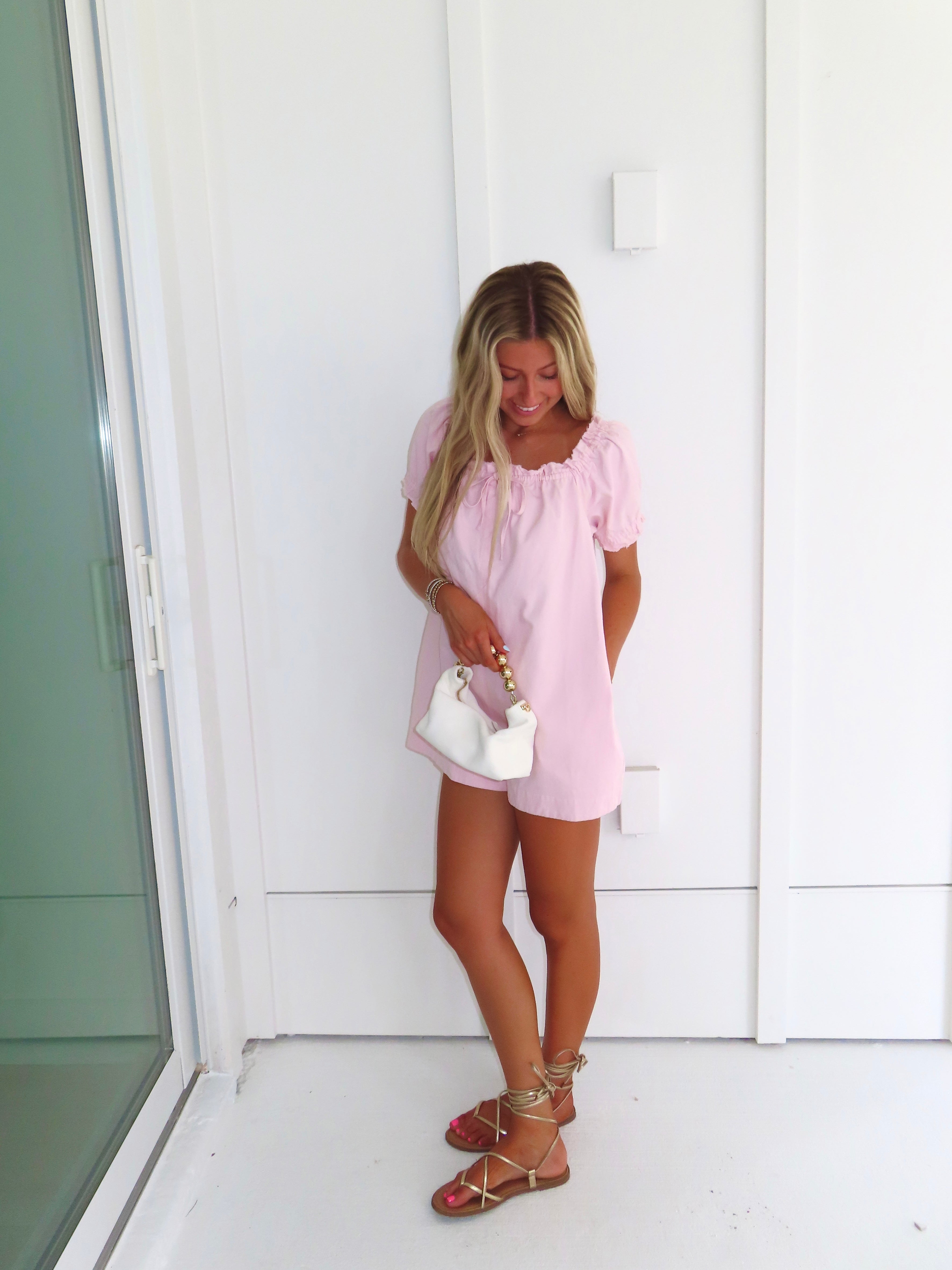 Woman wearing a pink romper and gold sandals and holding a white purse standing against a white wall.