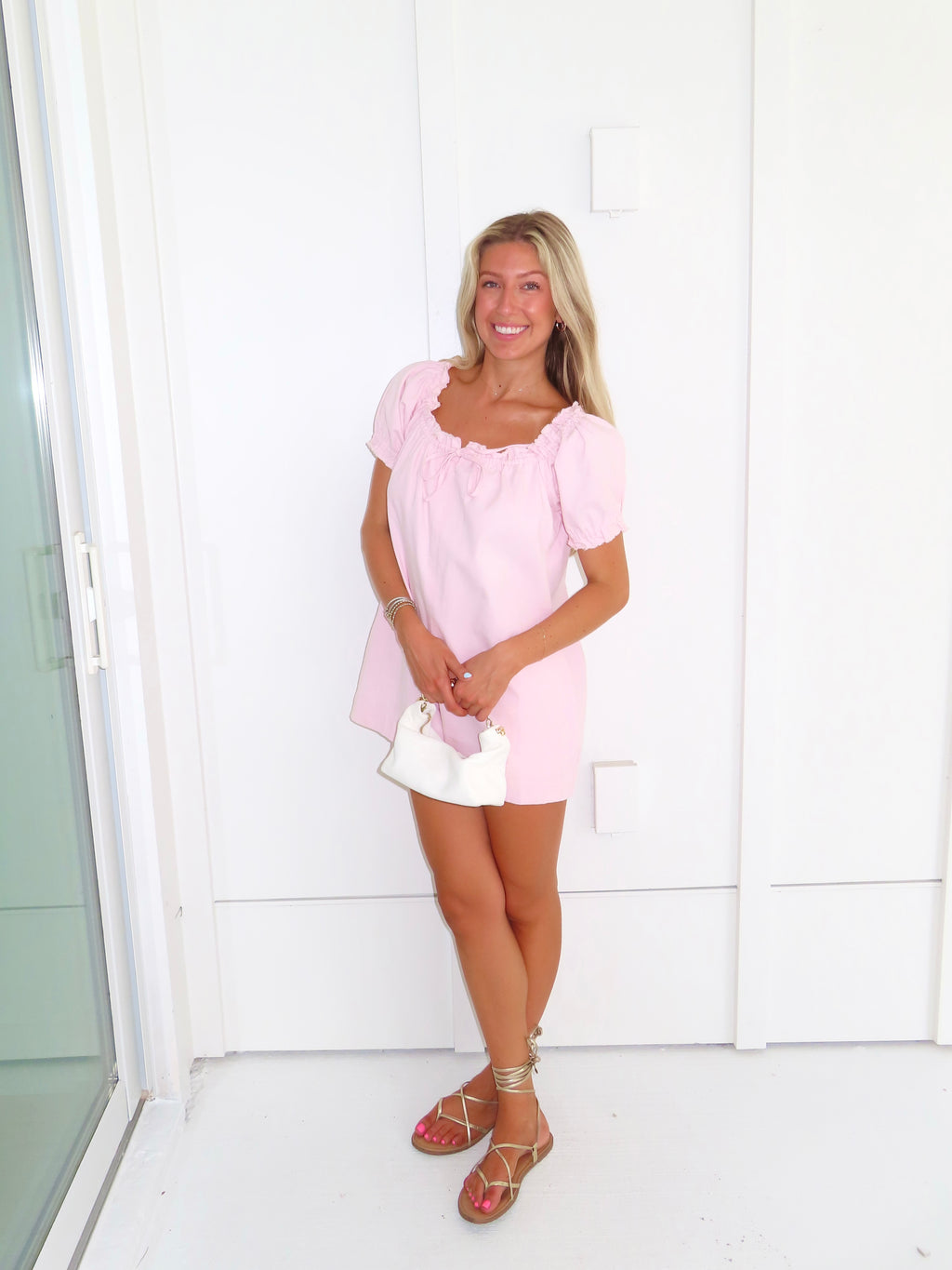 Woman wearing a pink romper and gold sandals and holding a white purse standing against a white wall.