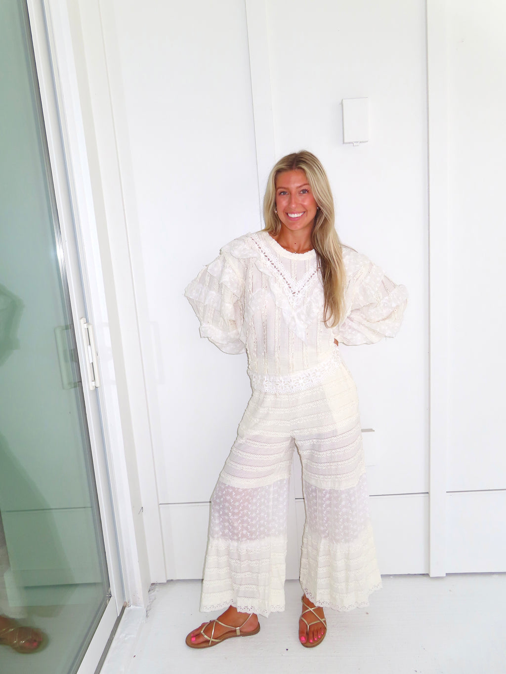 Woman standing against a white paneled wall wearing a cream lace detailed ruffle long sleeve top with matching pants and wearing gold sandals.