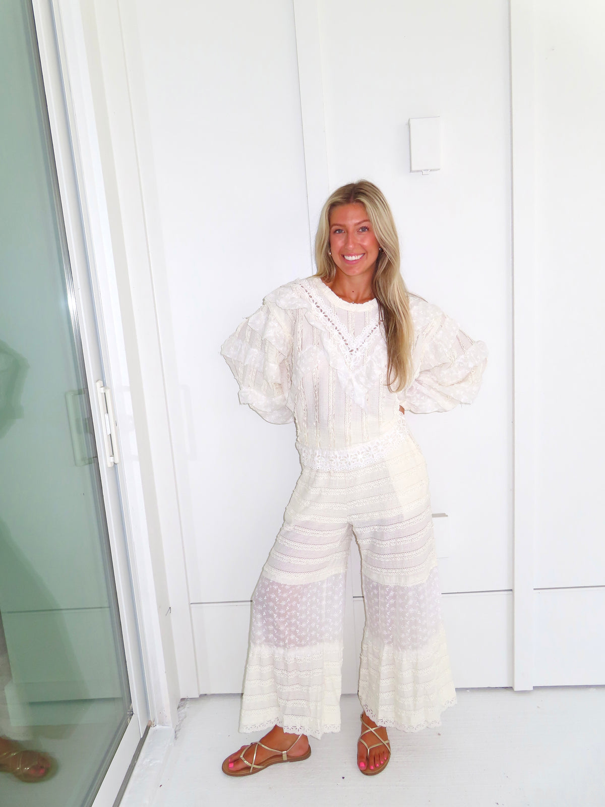 Woman standing against a white paneled wall wearing a cream lace detailed ruffle long sleeve top with matching pants and wearing gold sandals.