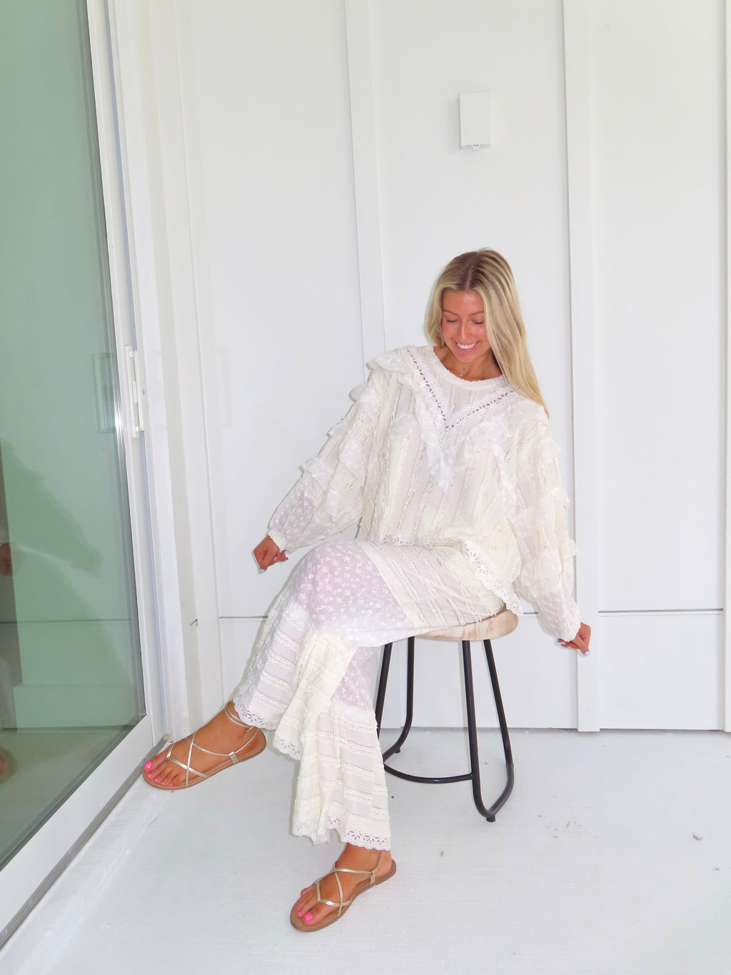 Woman sitting on a stool against a white paneled wall wearing a cream lace detailed ruffle long sleeve top with matching pants, wearing gold sandals.