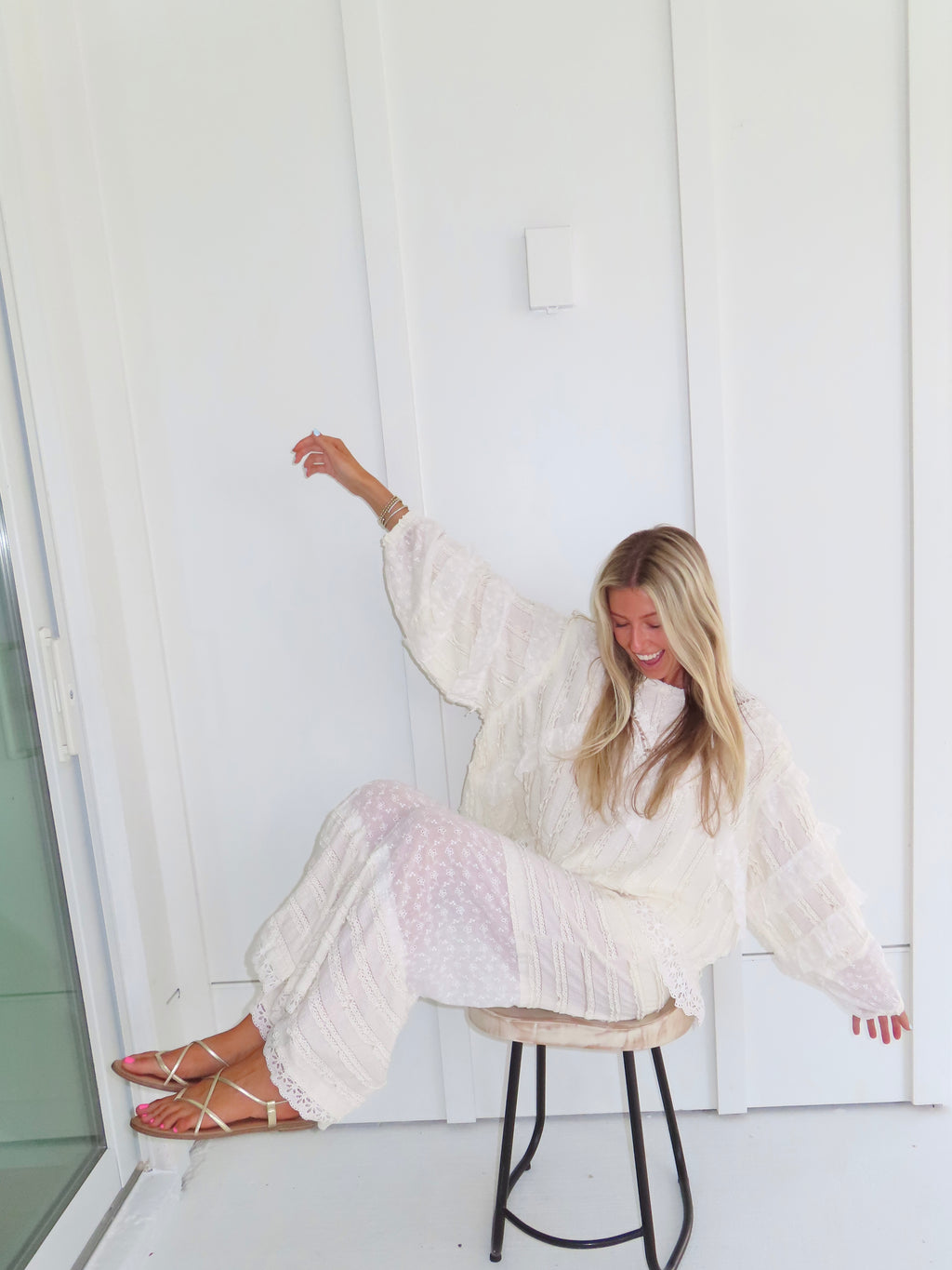 Woman sitting on a stool against a white paneled wall wearing a cream lace detailed long sleeve top with matching pants, wearing gold sandals and holding her arms up.
