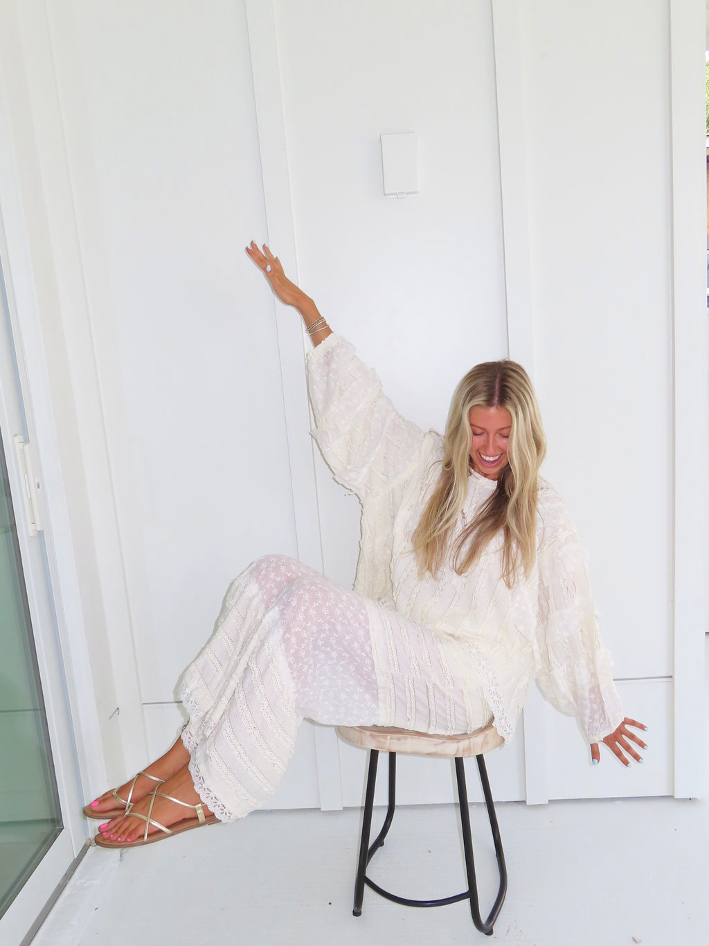Woman sitting on a stool against a white paneled wall wearing a cream lace detailed long sleeve top with matching pants, wearing gold sandals and holding her arms up.