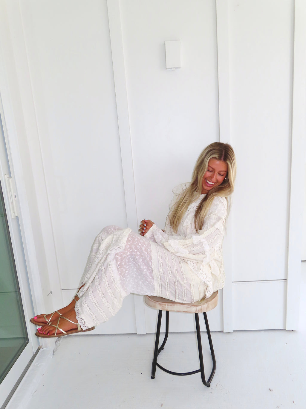 Woman sitting on a stool against a white paneled wall wearing a cream lace detailed ruffle long sleeve top with matching pants, wearing gold sandals with her legs up.