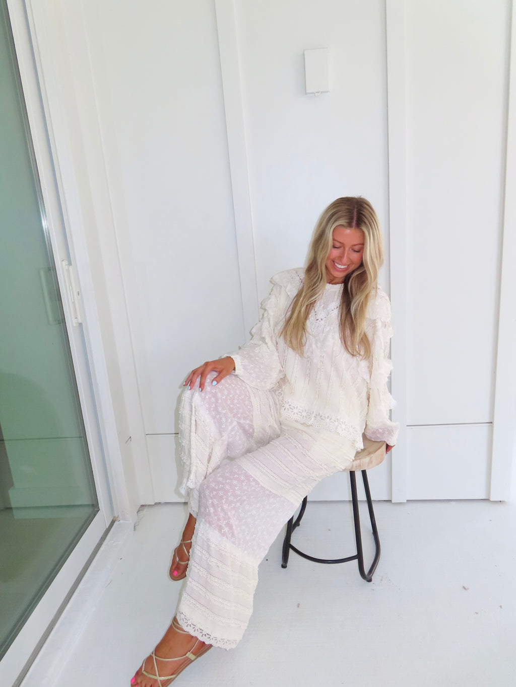 Woman sitting on a stool against a white paneled wall wearing a cream lace detailed long sleeve top with matching pants, wearing gold sandals.