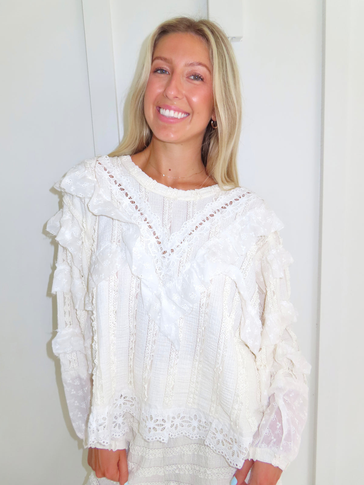 Woman standing against a white paneled wall wearing a cream lace detailed ruffle long sleeve top.