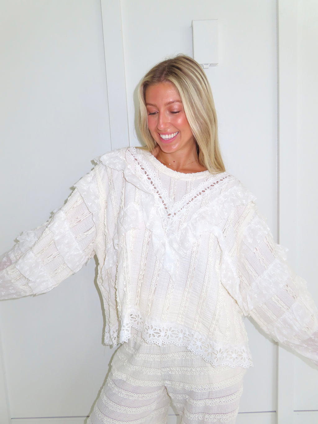 Woman standing against a white paneled wall wearing a cream lace detailed ruffle long sleeve top.