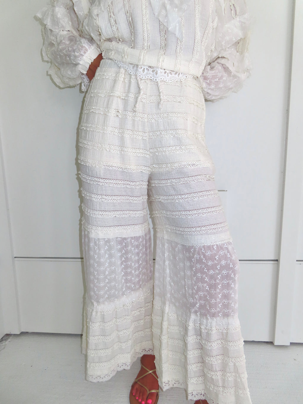 Woman standing against a white paneled wall wearing cream lace detailed ruffle pants with gold sandals.