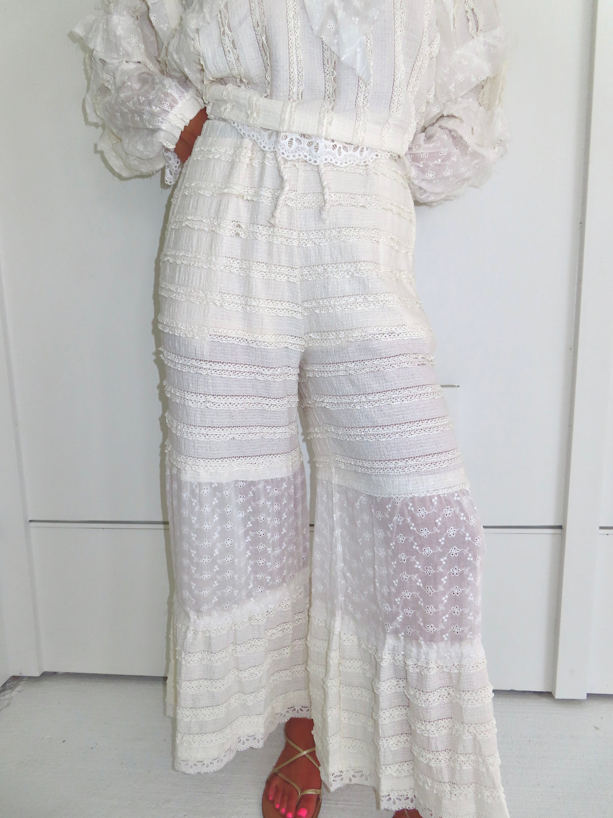 Woman standing against a white paneled wall wearing cream lace detailed ruffle pants with gold sandals.