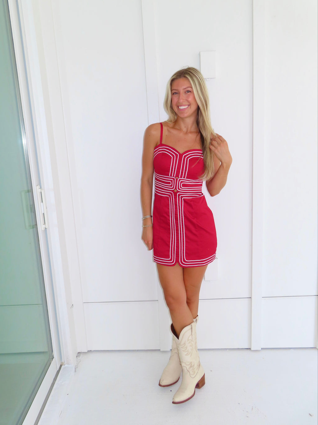 Woman wearing a red dress with white trim and cream cowboy boots standing against a white wall.
