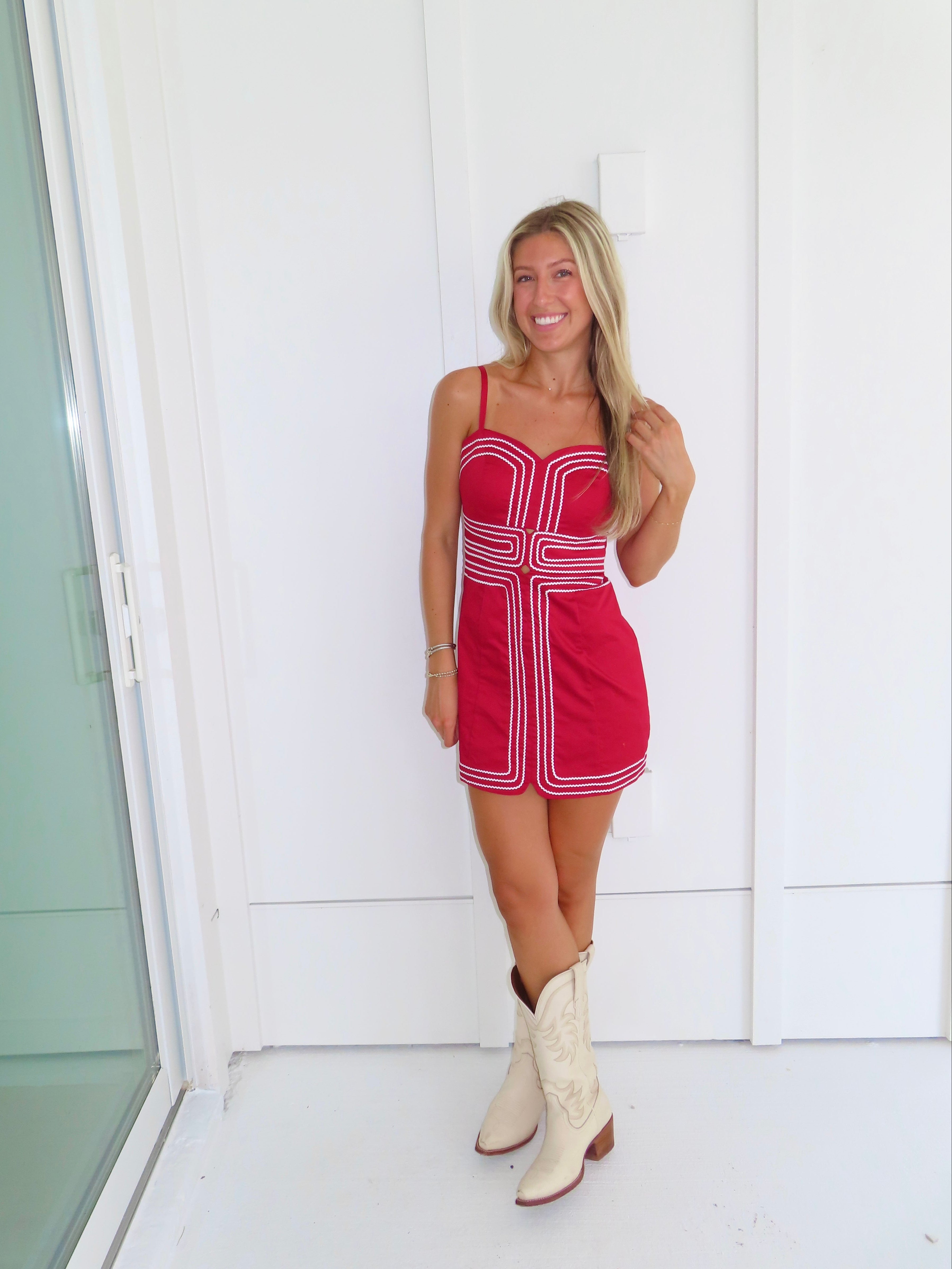 Woman wearing a red dress with white trim and cream cowboy boots standing against a white wall.