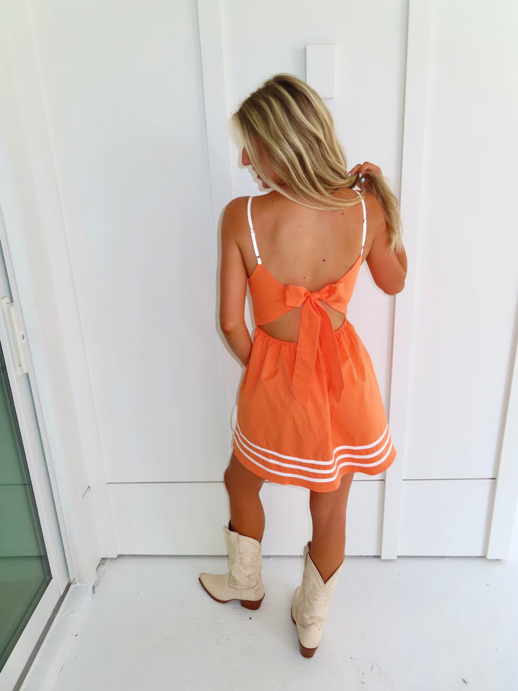A woman wearing a bright orange mini dress with white trim and cutout details, paired with cream cowboy boots and holding a small white handbag against a white wall.