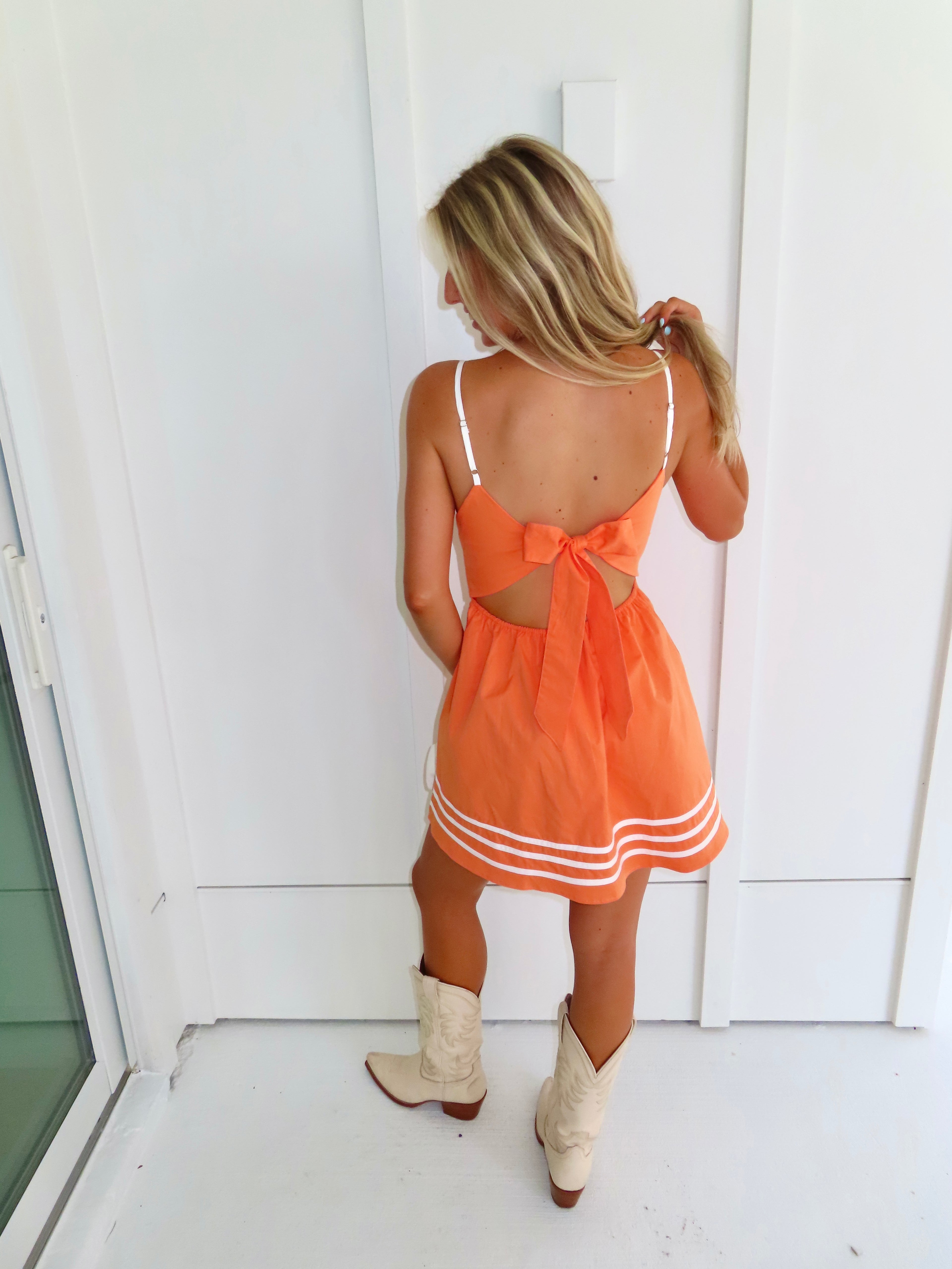 A woman wearing a bright orange mini dress with white trim and cutout details, paired with cream cowboy boots and holding a small white handbag against a white wall.