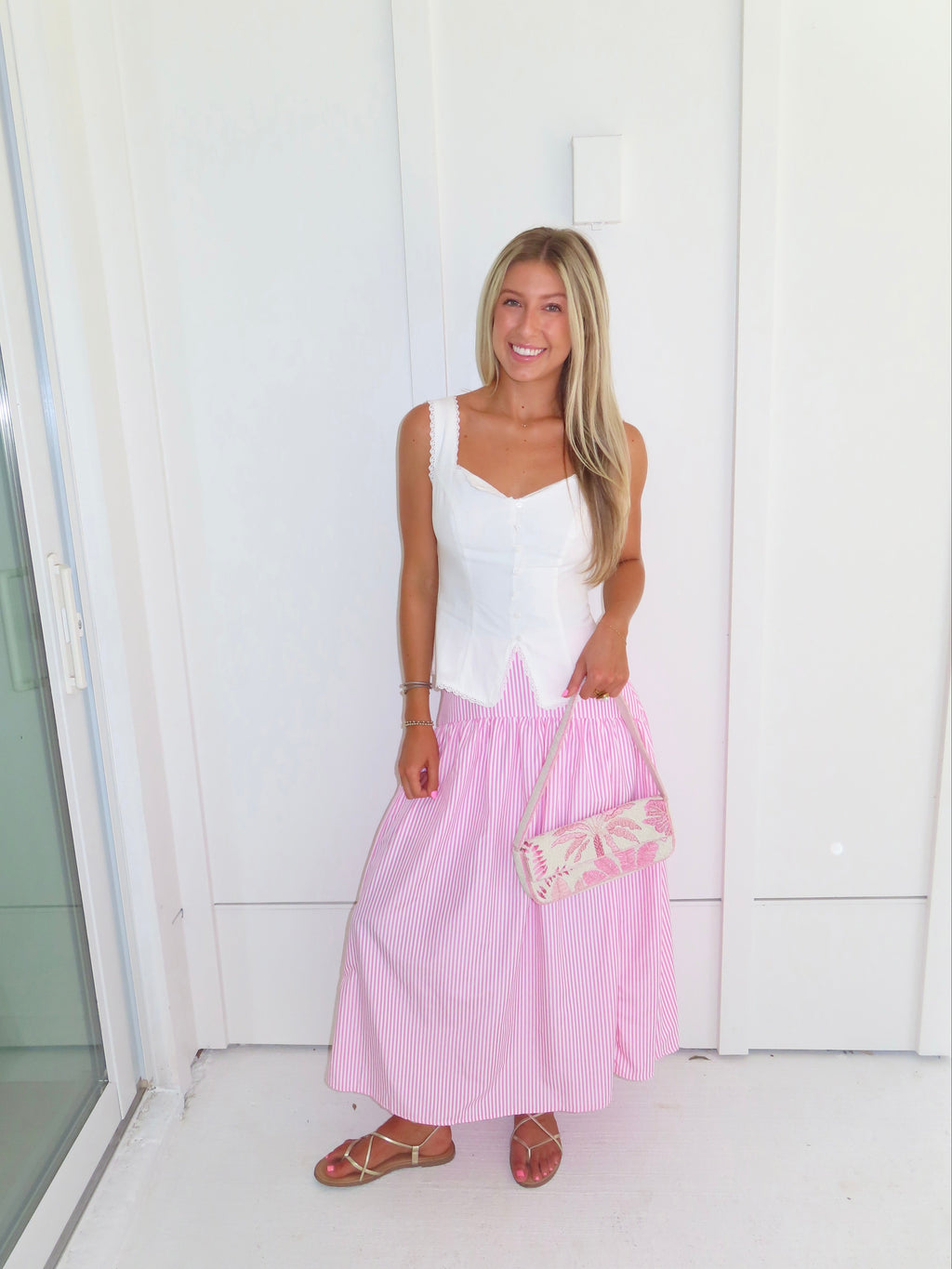 A woman wearing a pink and white striped maxi skirt with a white tank top and gold sandals, holding a pink and white purse and standing against a white paneled wall.